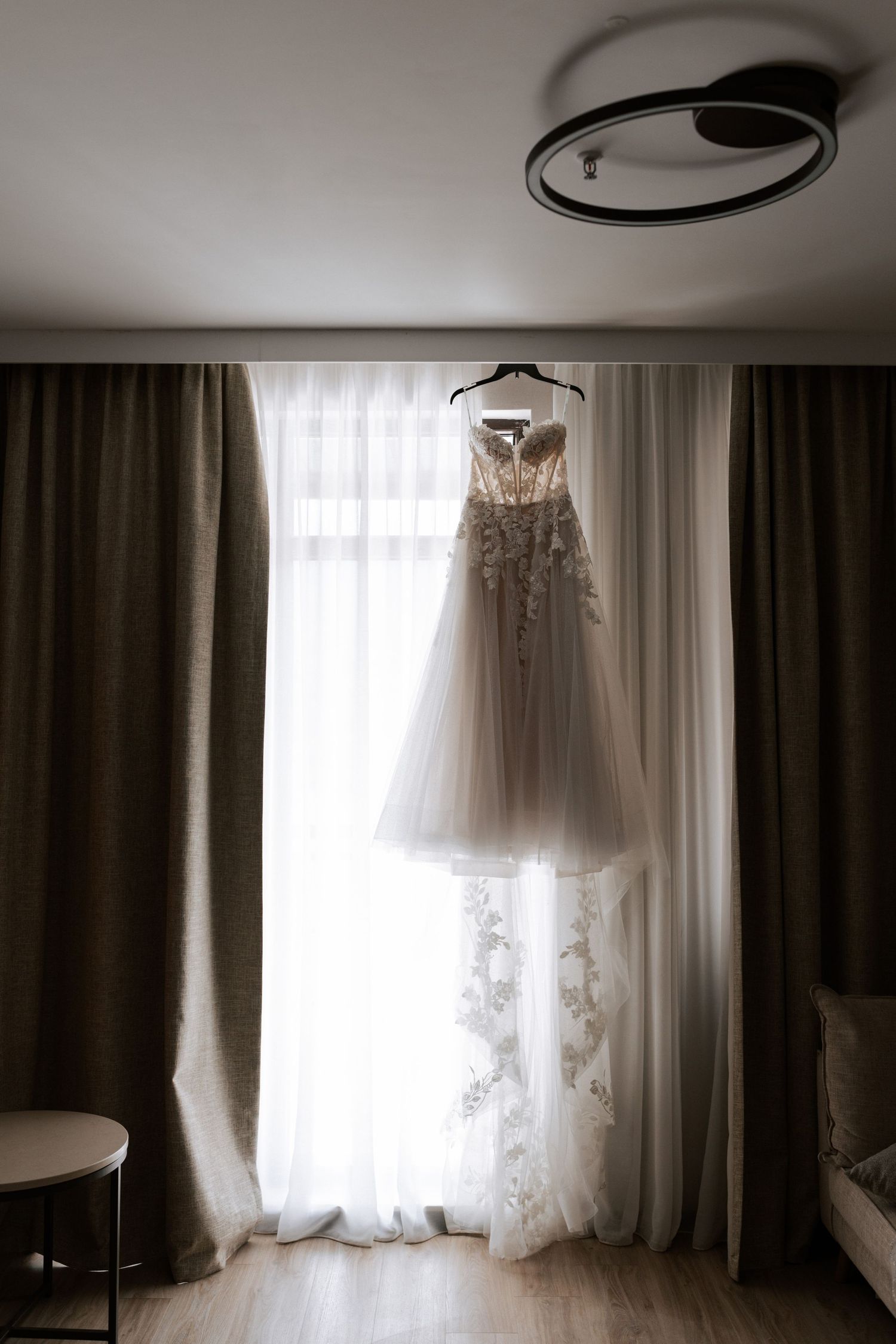 Elegant white lace wedding dress hanging in front of window with sheer curtains in natural light.