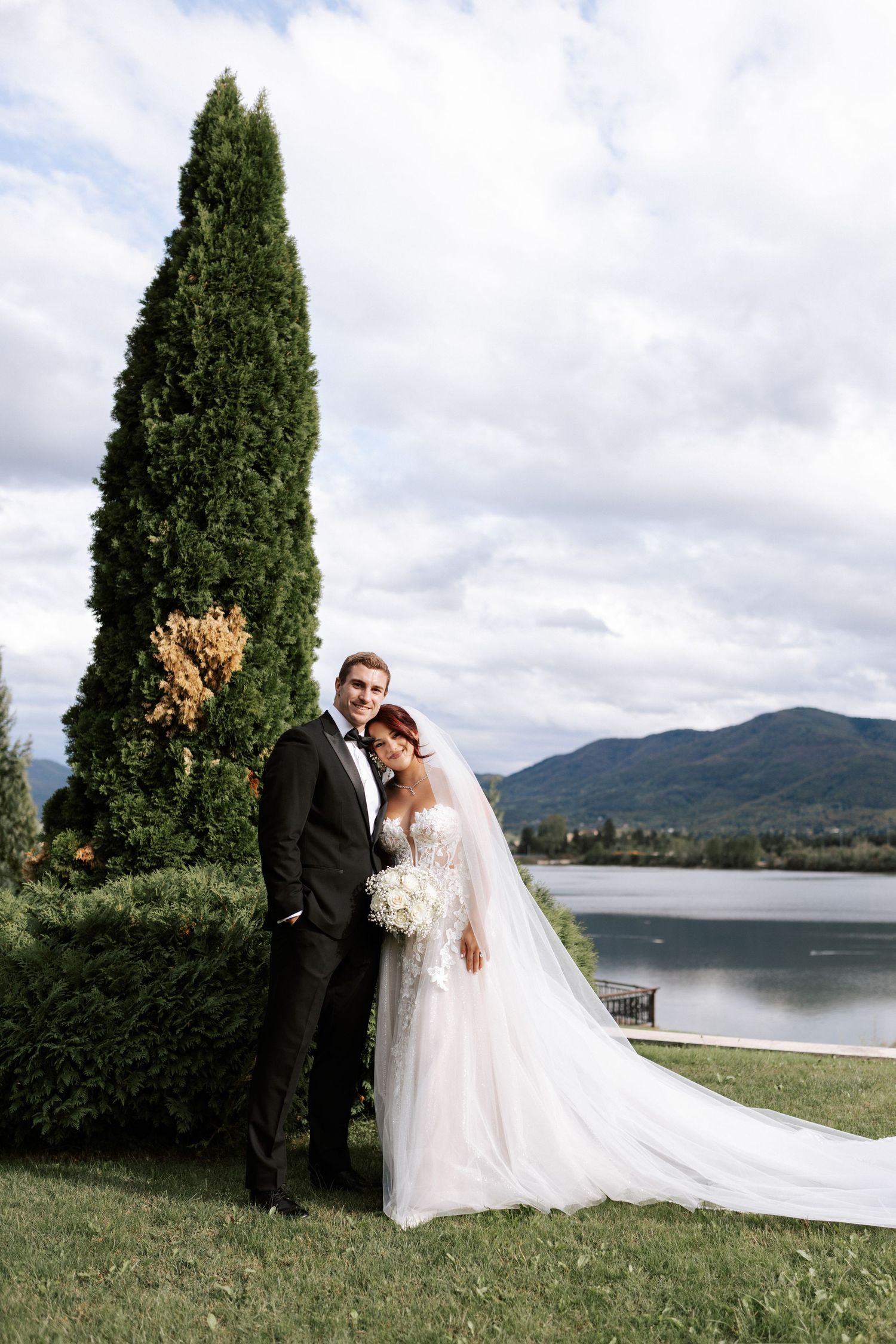 Wedding couple poses by tall cypress tree with lake and mountains in background.