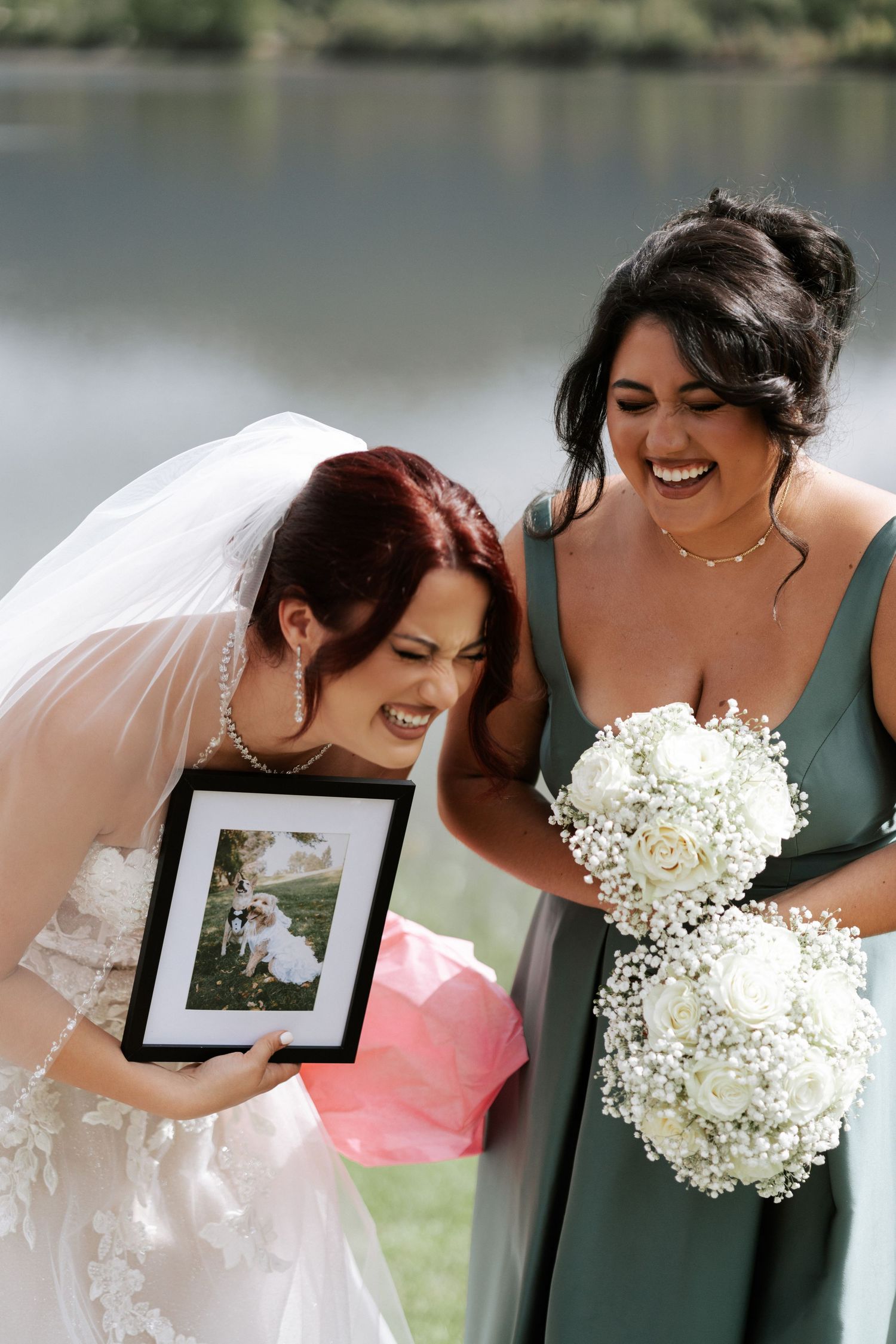 Two women share a joyful moment looking at a framed photo during a wedding ceremony by a lake.
