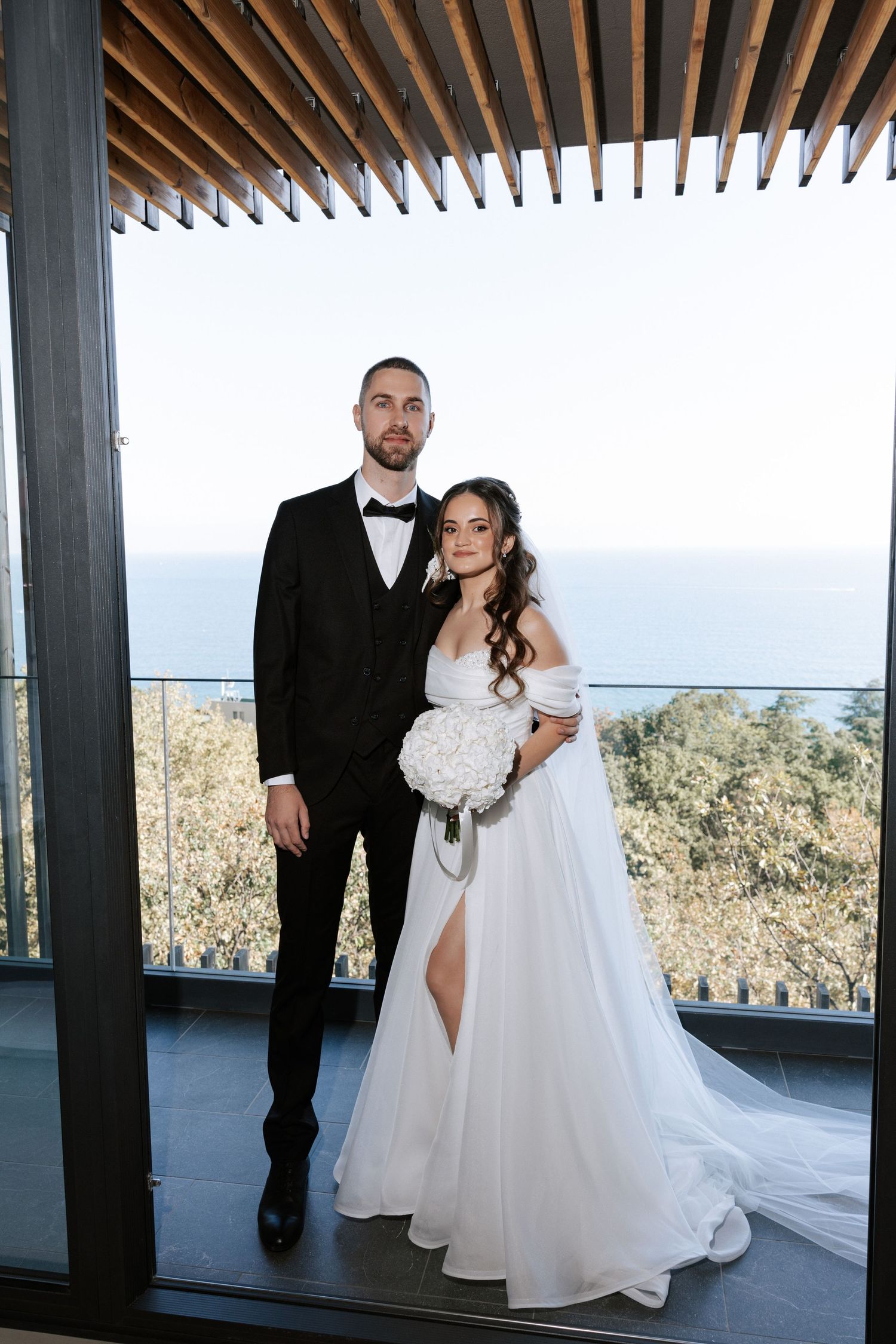 Wedding couple poses beneath wooden beams on a modern balcony overlooking the sea.