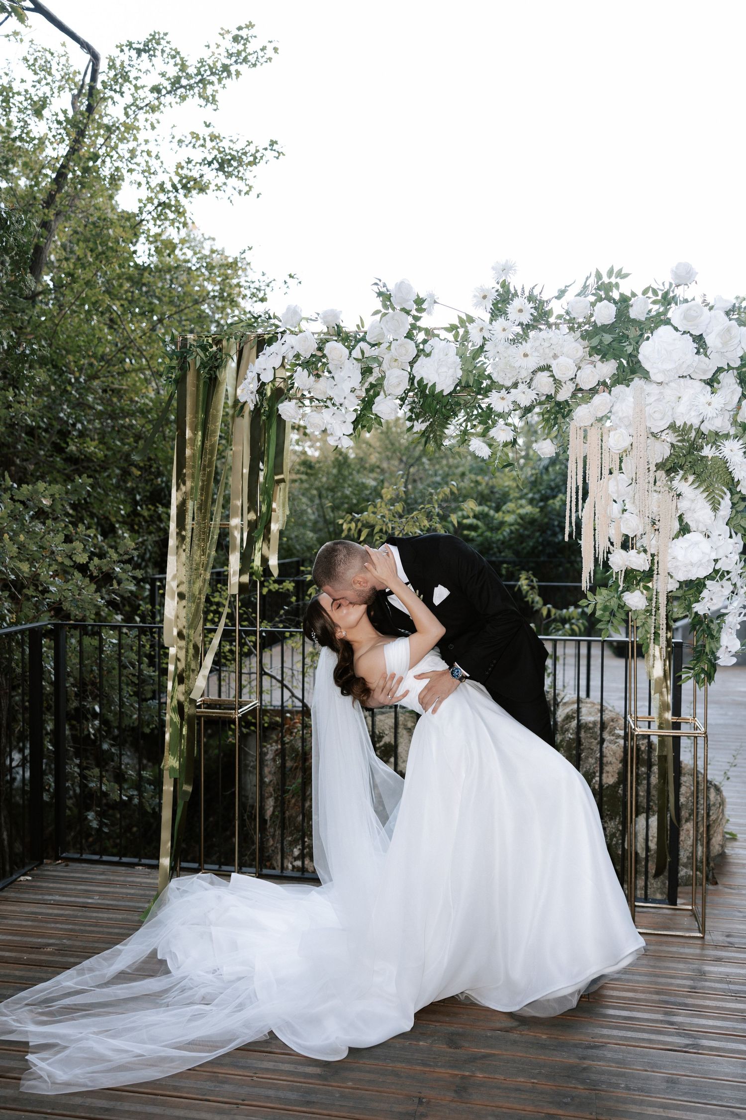 Romantic dip pose during wedding photoshoot under white floral arch outdoors.