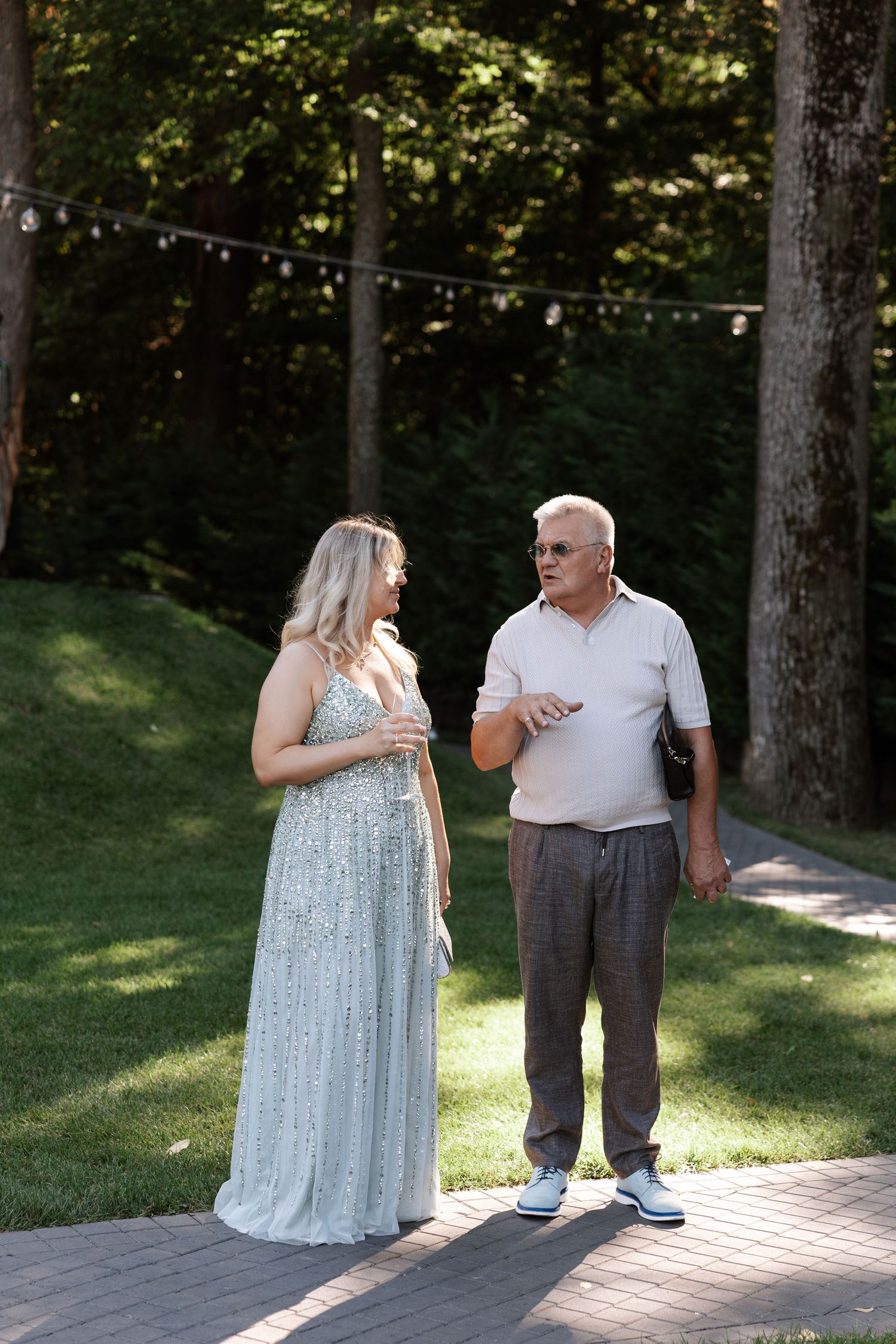 Couple walks and talks on brick pathway surrounded by trees in outdoor garden setting.