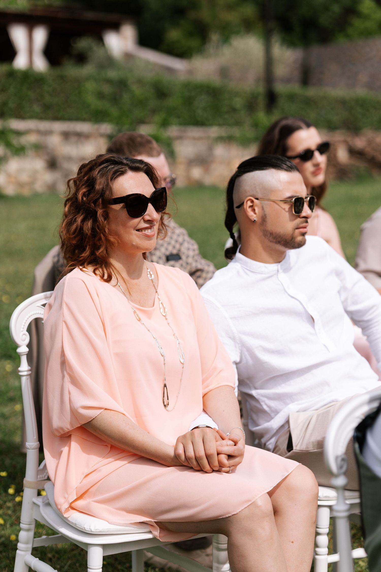 A couple in white and peach colored outfits sit together in white chairs outdoors, laughing and enjoying the sunshine.