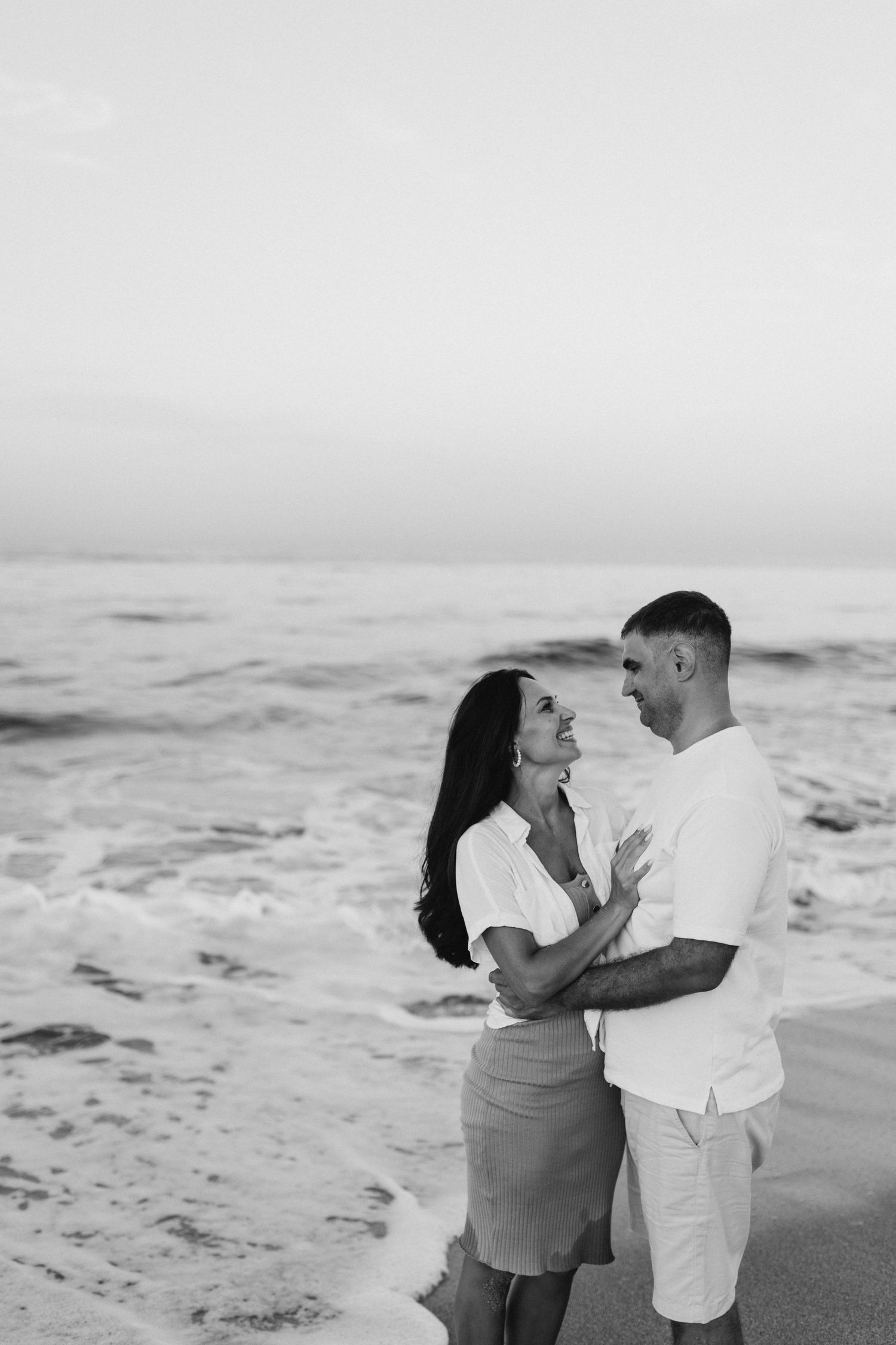 Black and white photo of a romantic couple embracing on a beach with ocean waves behind them.