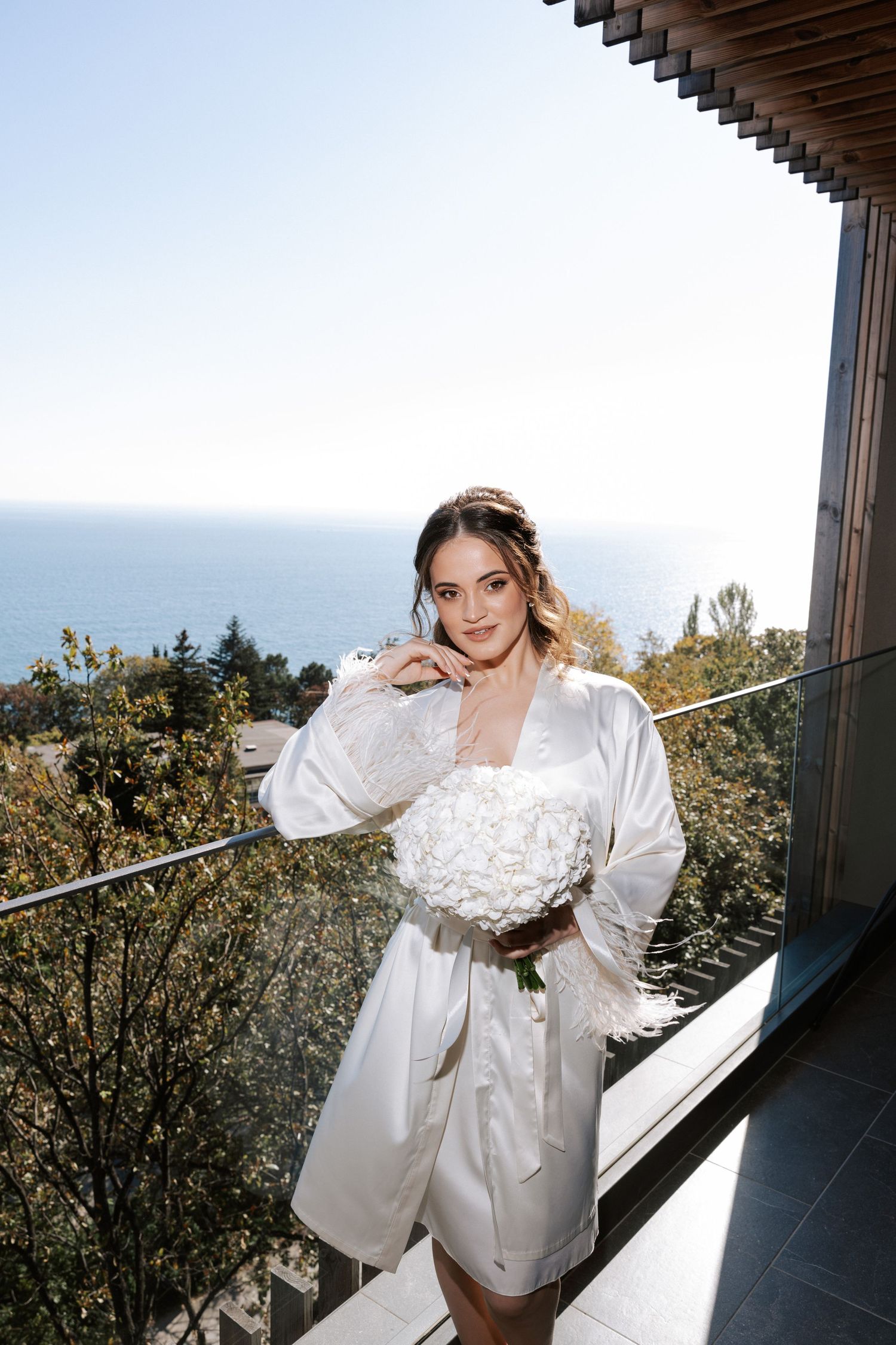 A bride in a white silk robe poses on a balcony overlooking the ocean view.