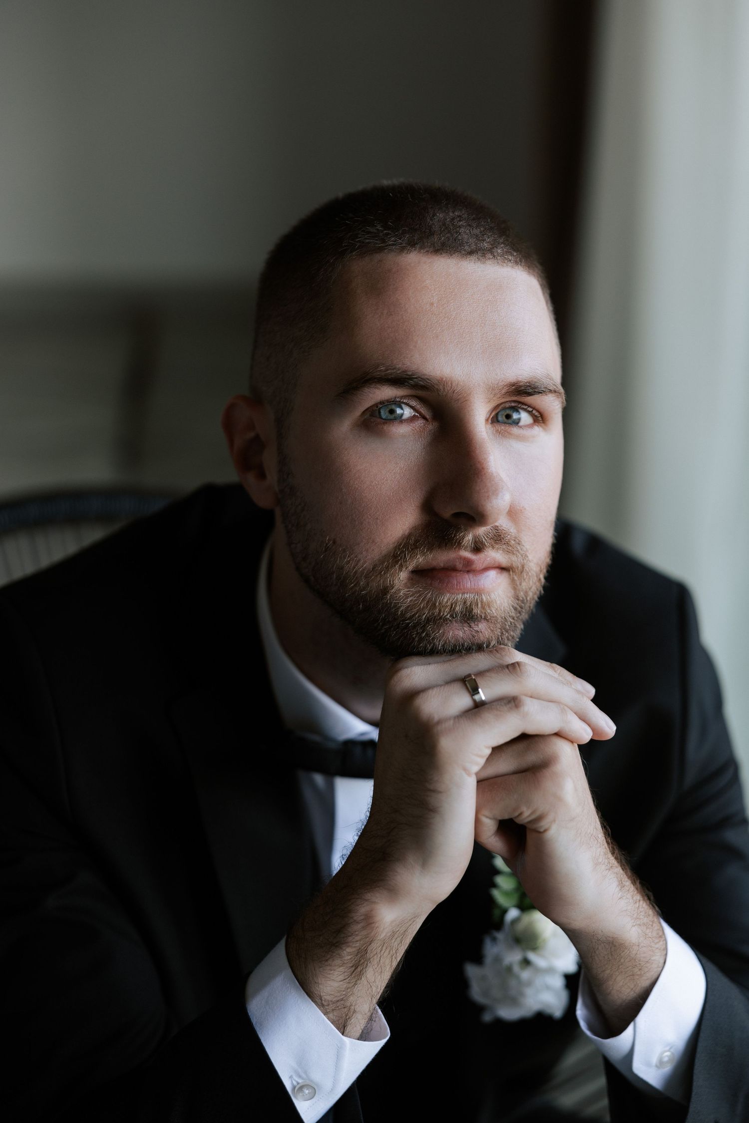 Groom in black tuxedo and bowtie poses thoughtfully in window light.