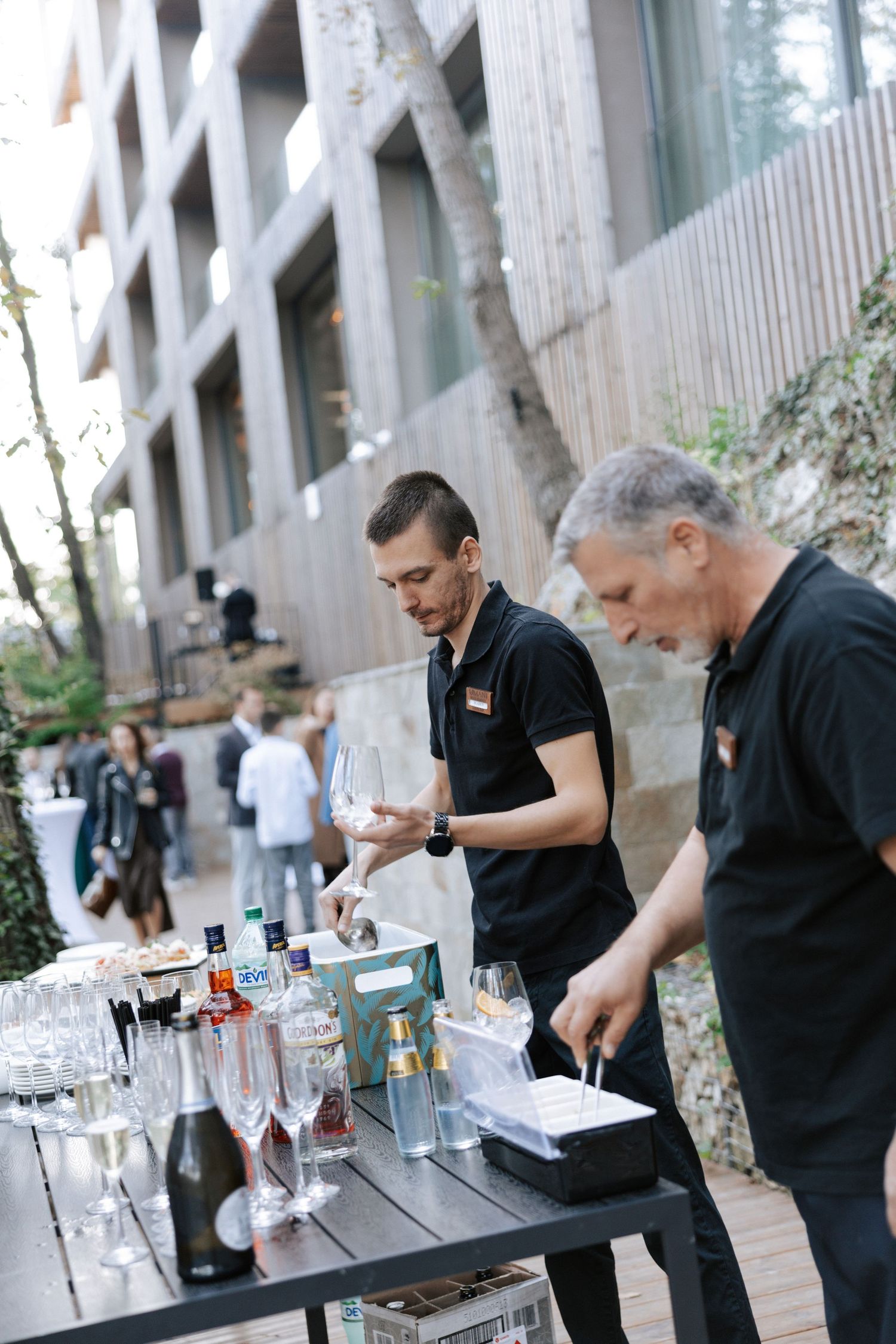Bartenders preparing drinks at outdoor wedding reception near modern building.