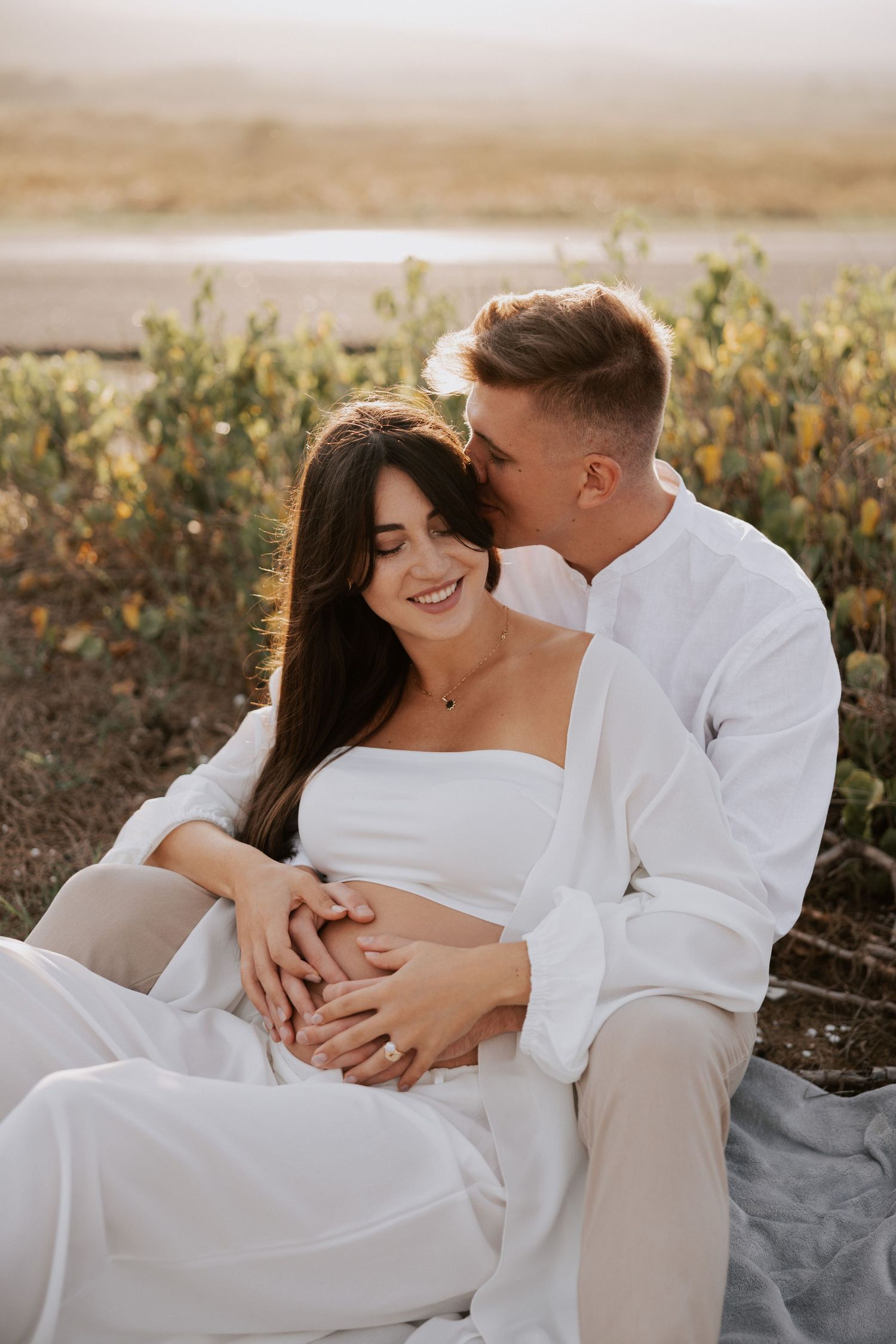 Romantic sunset photoshoot of couple in white attire enjoying intimate moment outdoors.