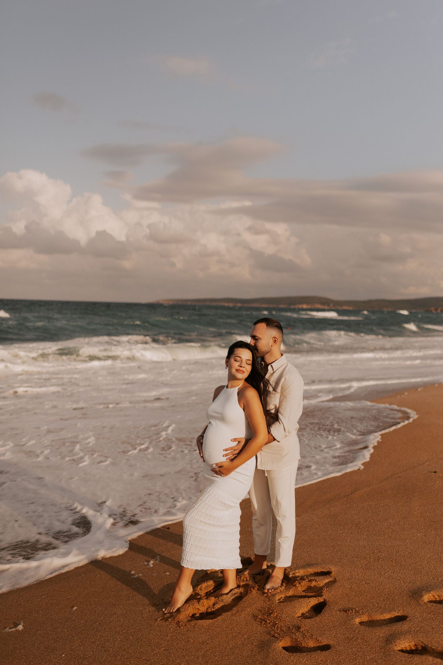 A couple embraces on a beach at sunset with crashing waves and cloudy skies in the background.