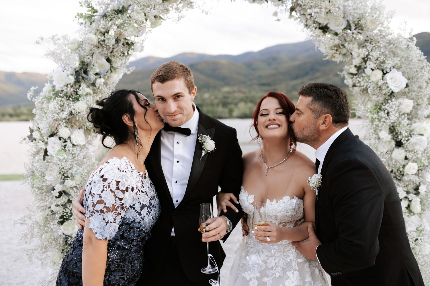 Wedding party shares laughs under white floral arch with mountain views.