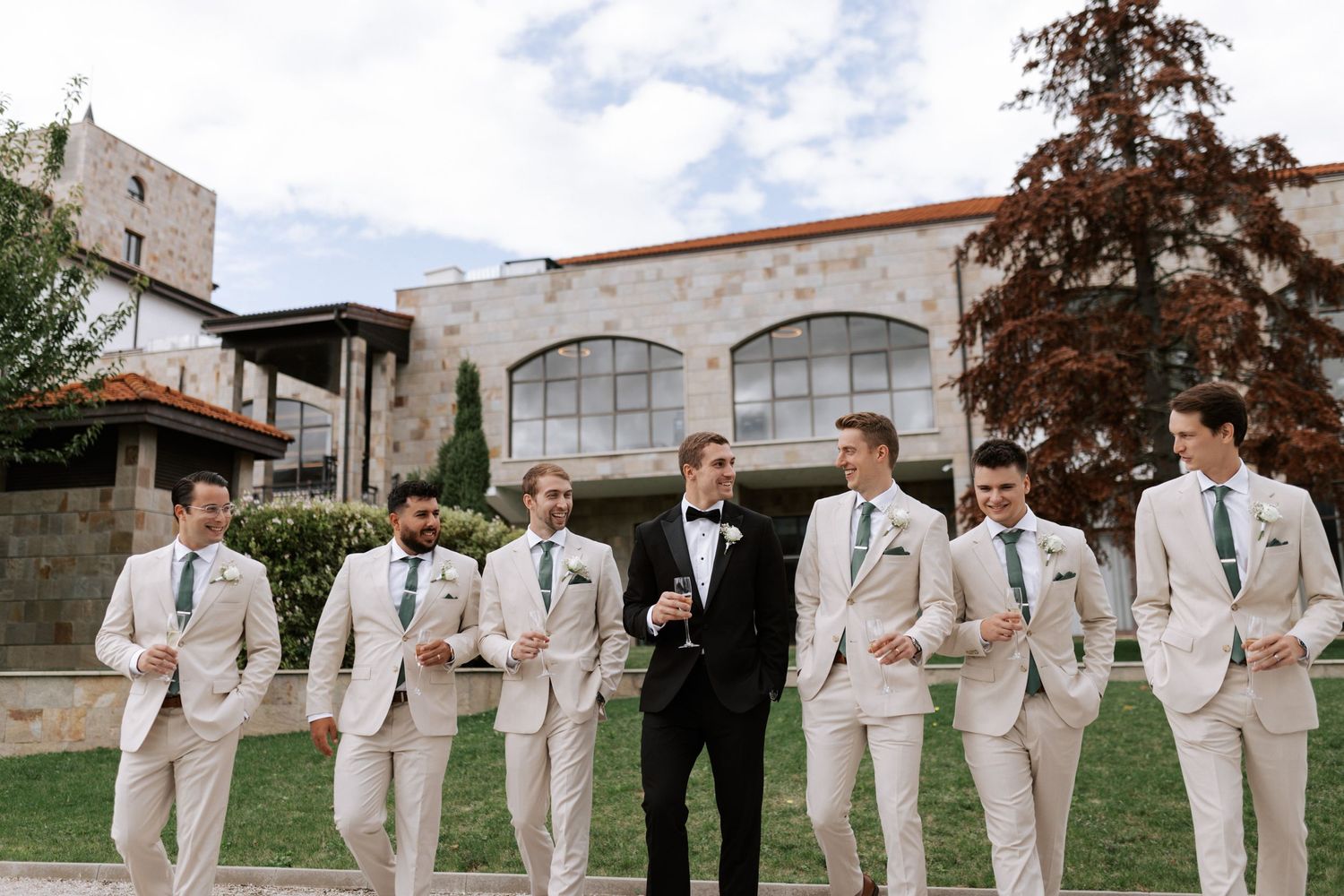 Groomsmen in light beige suits pose with one in black at a stone building with arched windows.