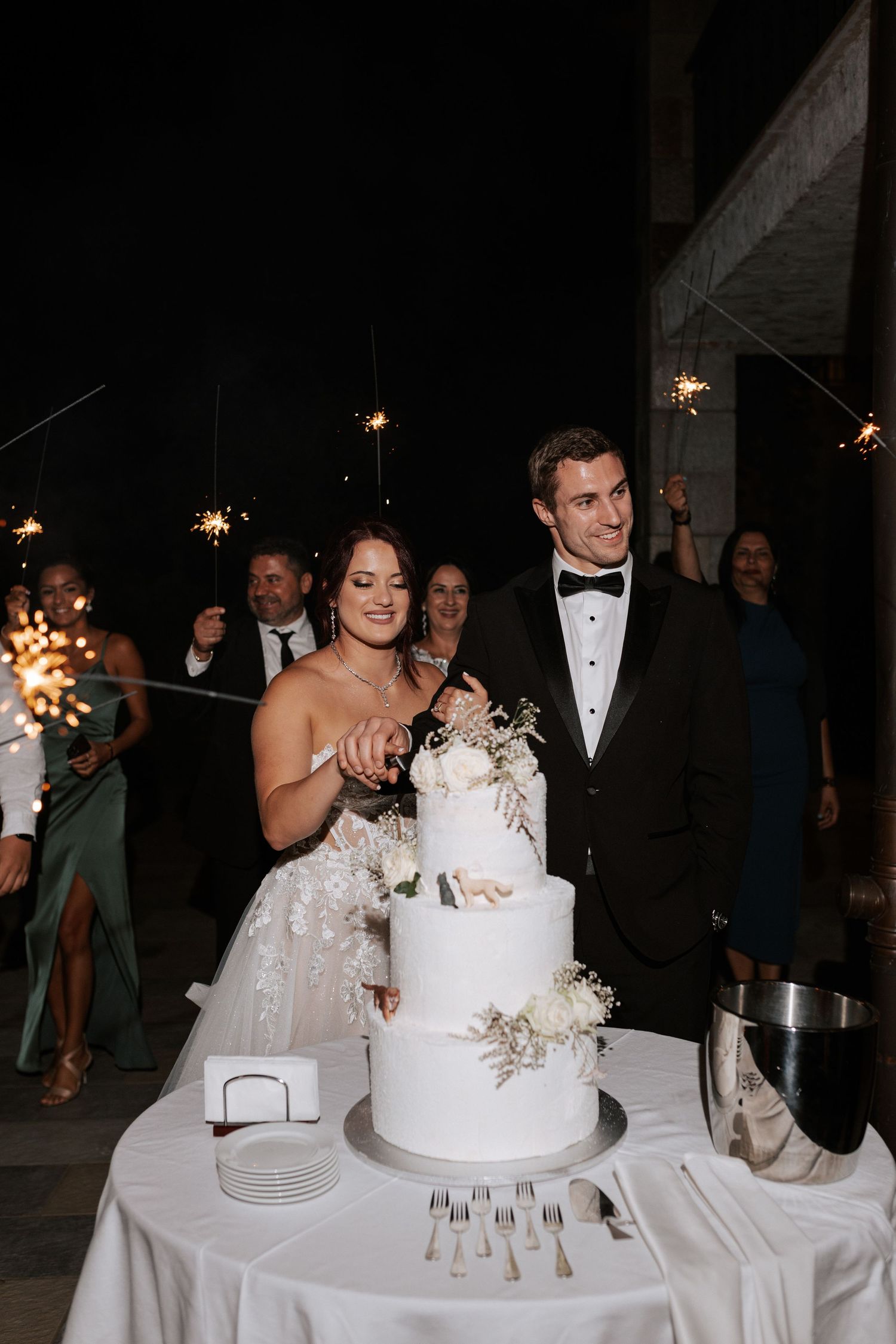 Wedding couple stands by their elegant white tiered wedding cake decorated with delicate floral details.