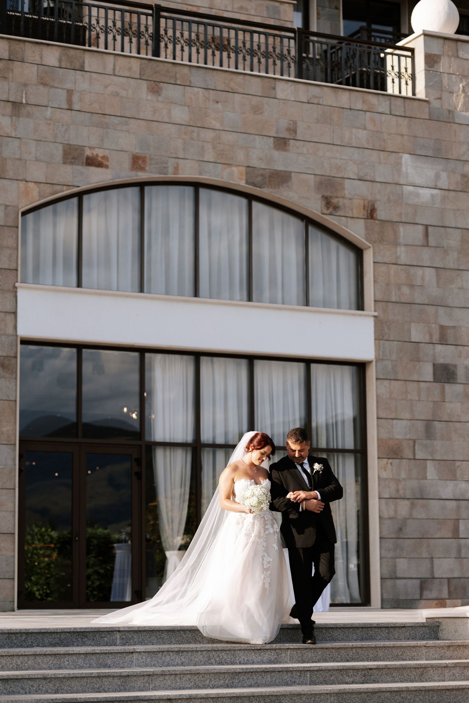 A couple exits down stone steps outside a building with arched windows.