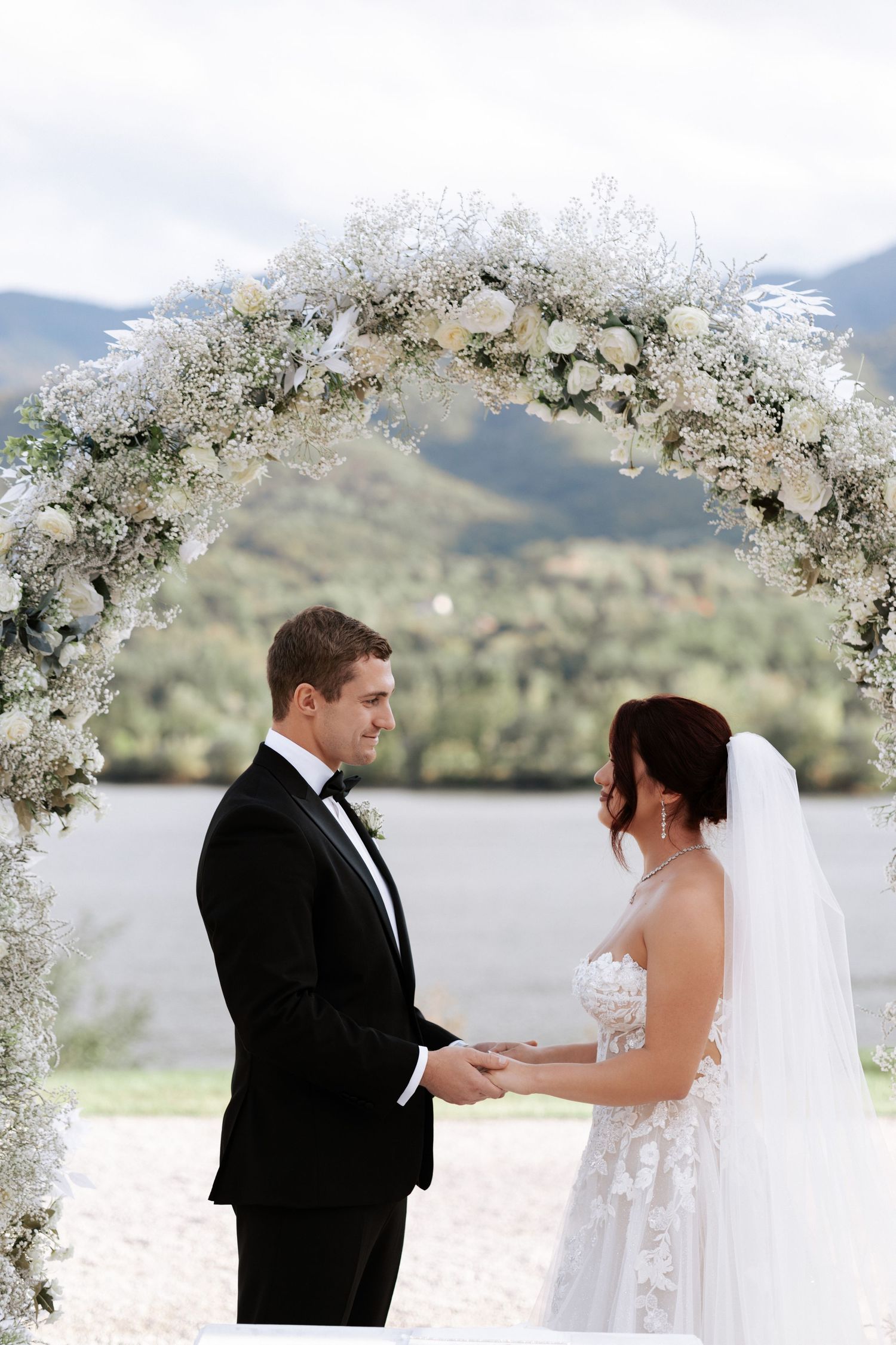A couple exchanges vows beneath an elegant white floral arch with mountain scenery.