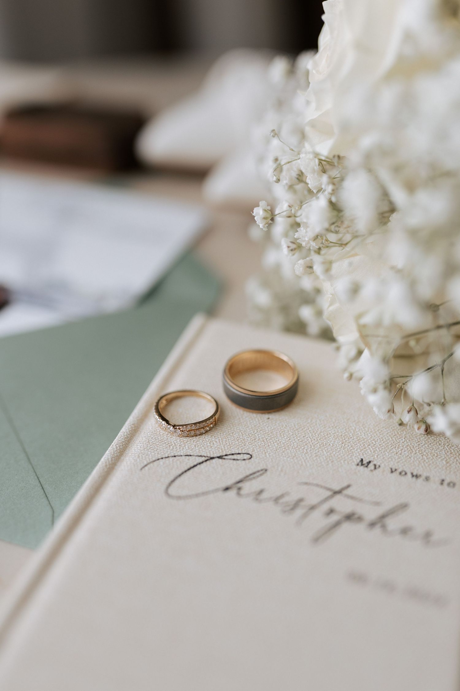 Gold wedding rings displayed on vow book with baby's breath flowers and sage green envelope.