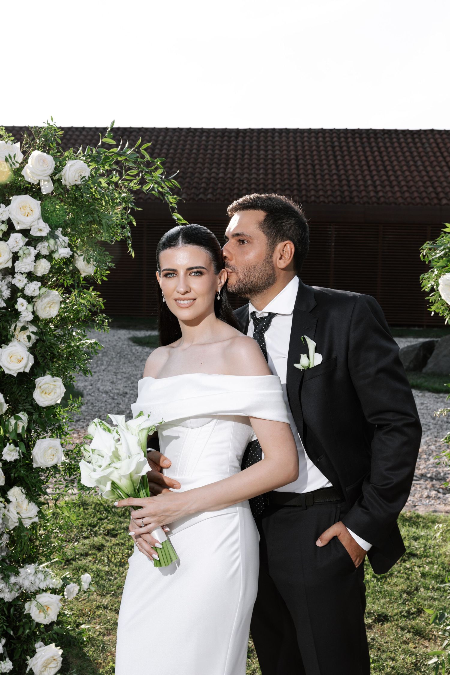 A romantic wedding couple embraces outdoors surrounded by white floral decorations and greenery.