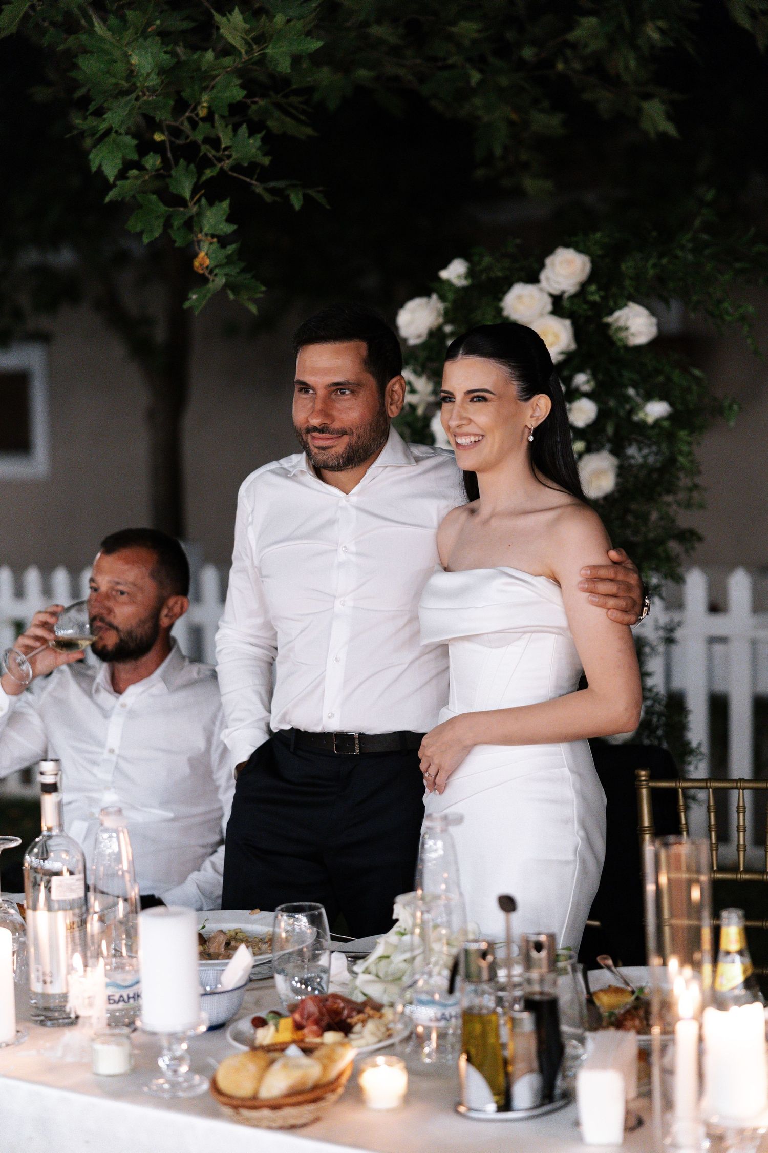 Wedding party guests share joyful moments at an elegant outdoor reception dinner table with candlelight and white decor.