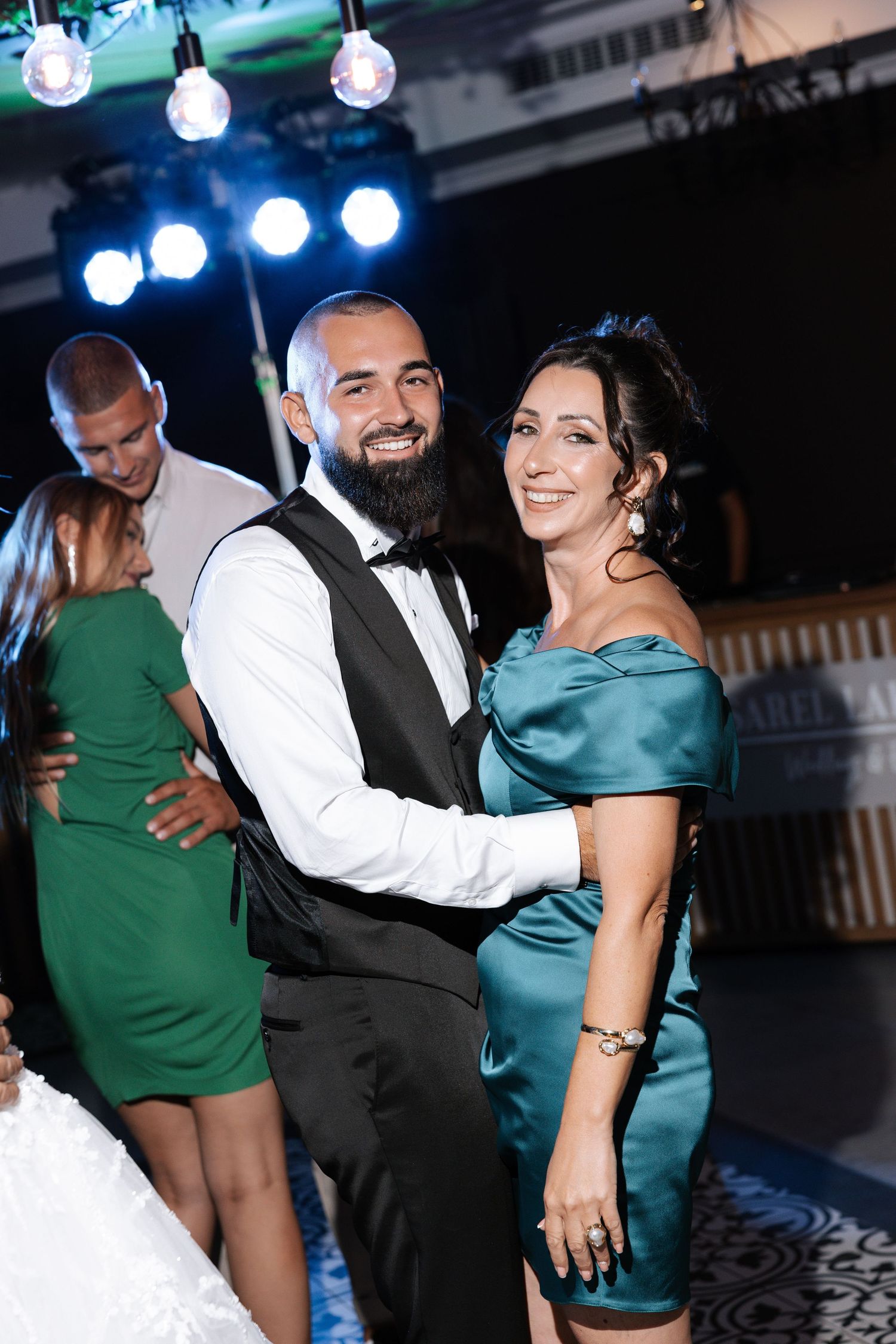 A couple in formal attire dancing at an evening event with blue lighting and decorative bulbs overhead.