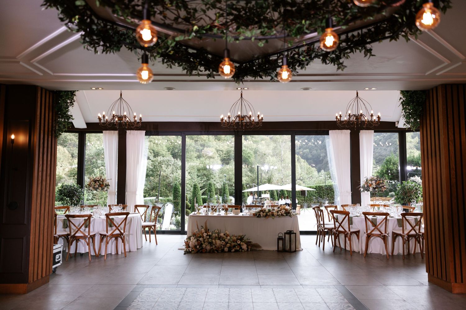 Elegant wedding reception venue with string lights, chandeliers, and floor-to-ceiling windows overlooking lush gardens.