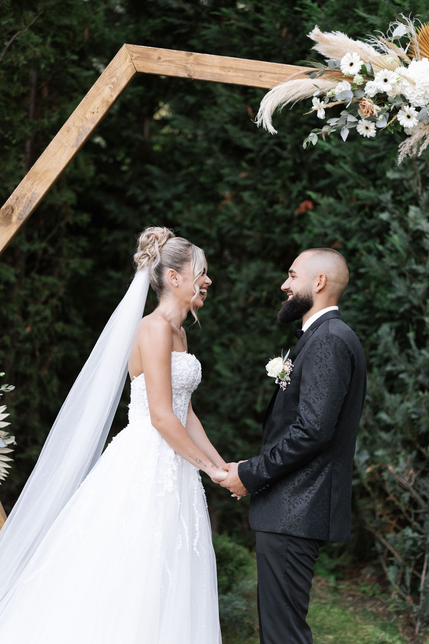 A romantic outdoor wedding ceremony shows a couple sharing a joyful moment under a wooden arch decorated with white flowers.