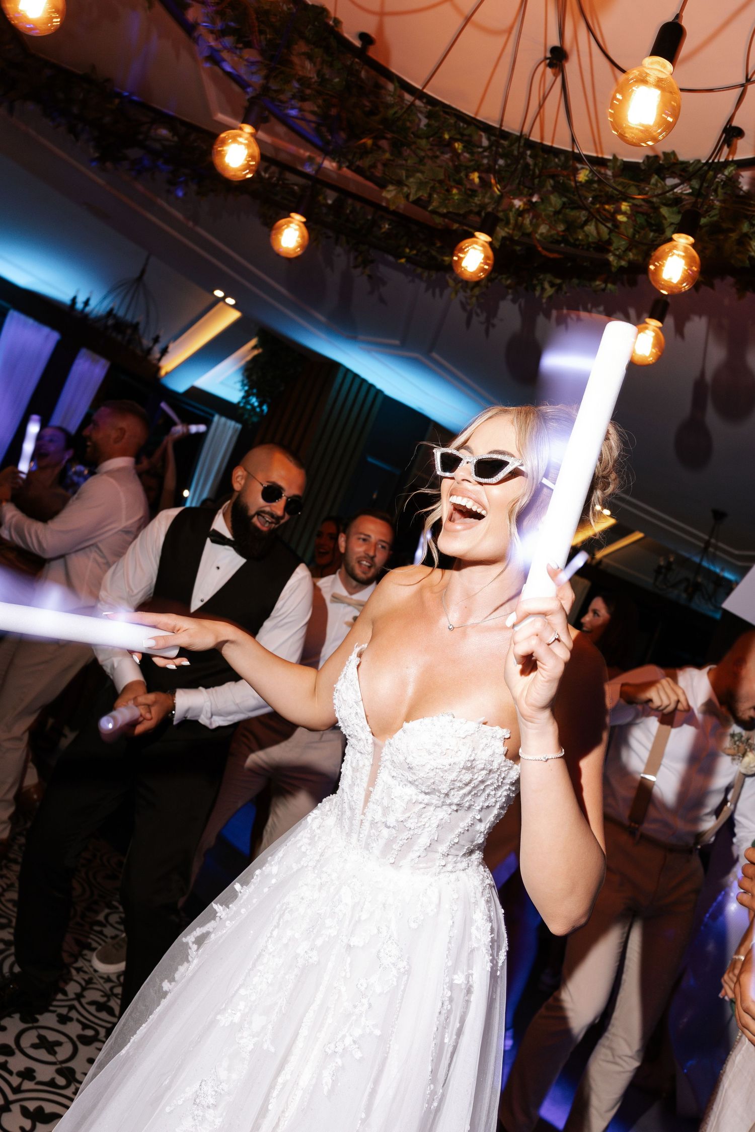Wedding reception guests dancing and celebrating under string lights and greenery decorations at an indoor venue.