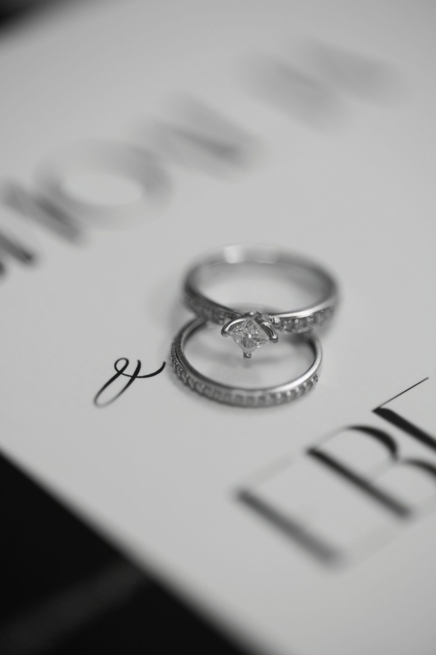A close-up black and white photograph of an engagement ring and wedding band on a textured surface.