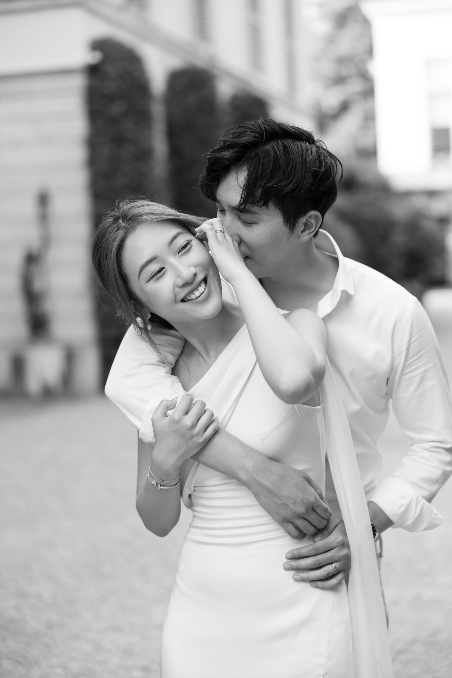 A romantic black and white portrait of a couple in white outfits embracing and laughing together outdoors.