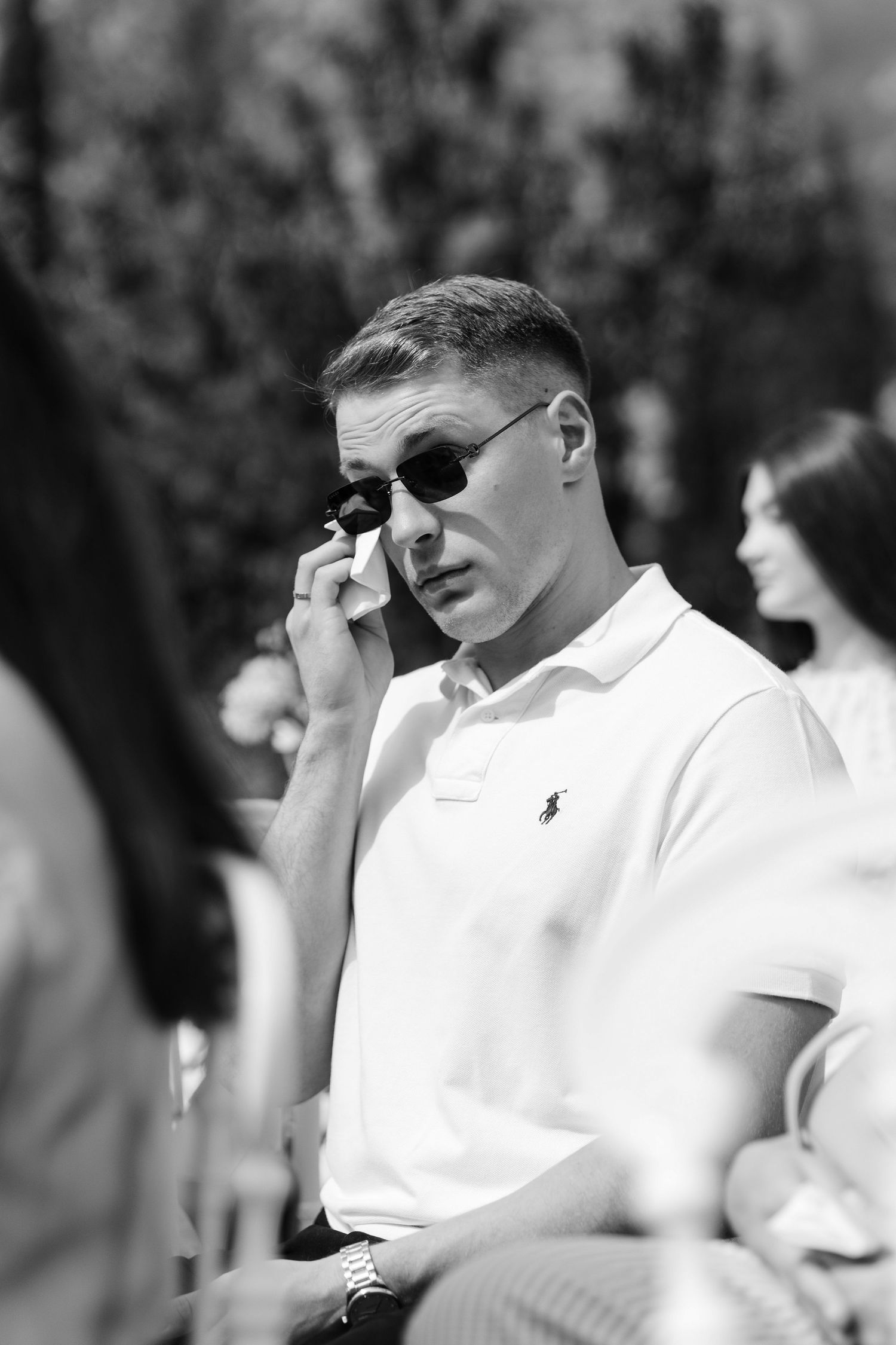 A person wearing a white polo shirt and sunglasses sits outdoors in black and white photography.