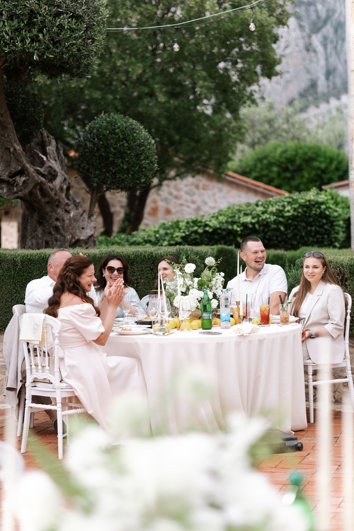 Guests seated at elegant outdoor wedding reception tables with white linens under trees in a garden setting.