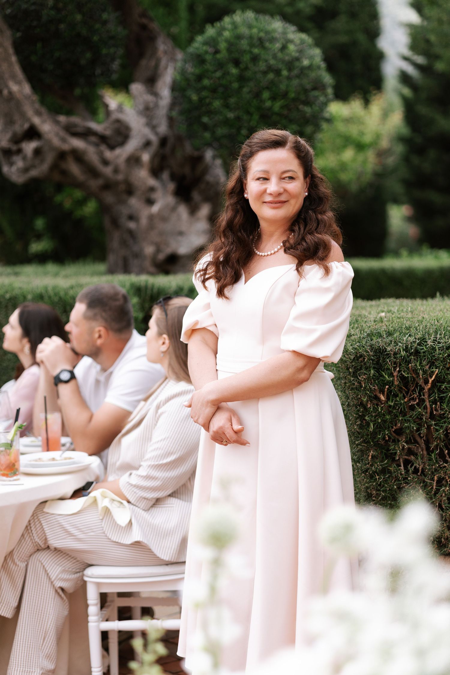 Family wearing white and blush outfits sits together on bench in garden setting for outdoor photo session.