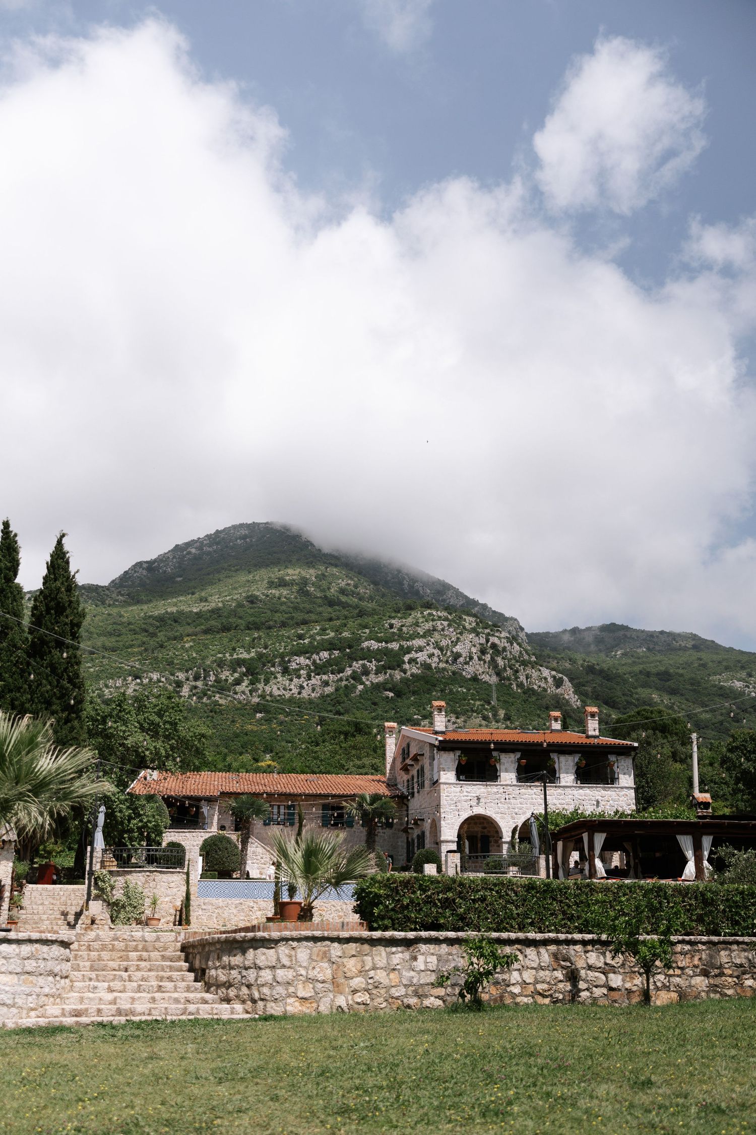 Mediterranean villa with stone walls and terracotta roof nestled against green mountain landscape under cloudy sky.