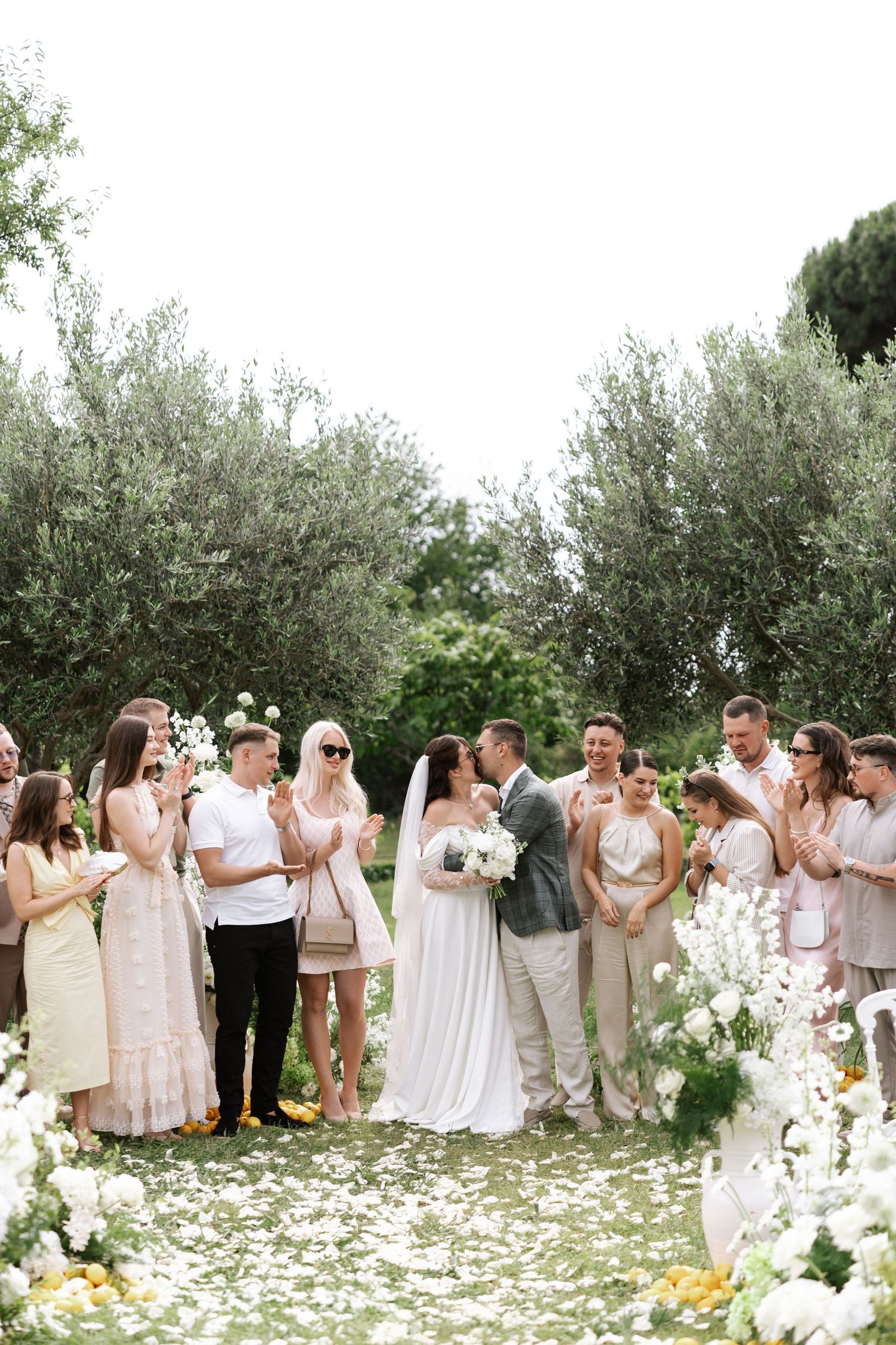 Wedding party poses in a line outdoors with olive trees and scattered white flower petals creating a romantic backdrop.