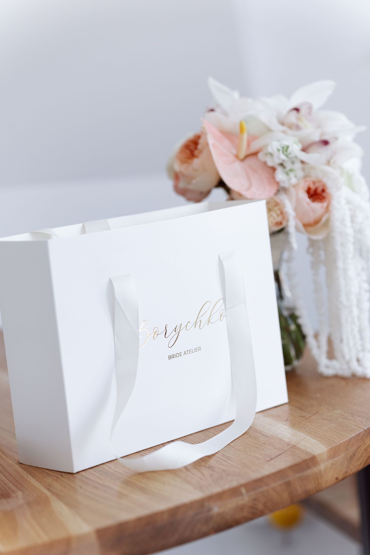 White shopping bag with gold lettering next to delicate pink and white floral arrangement.