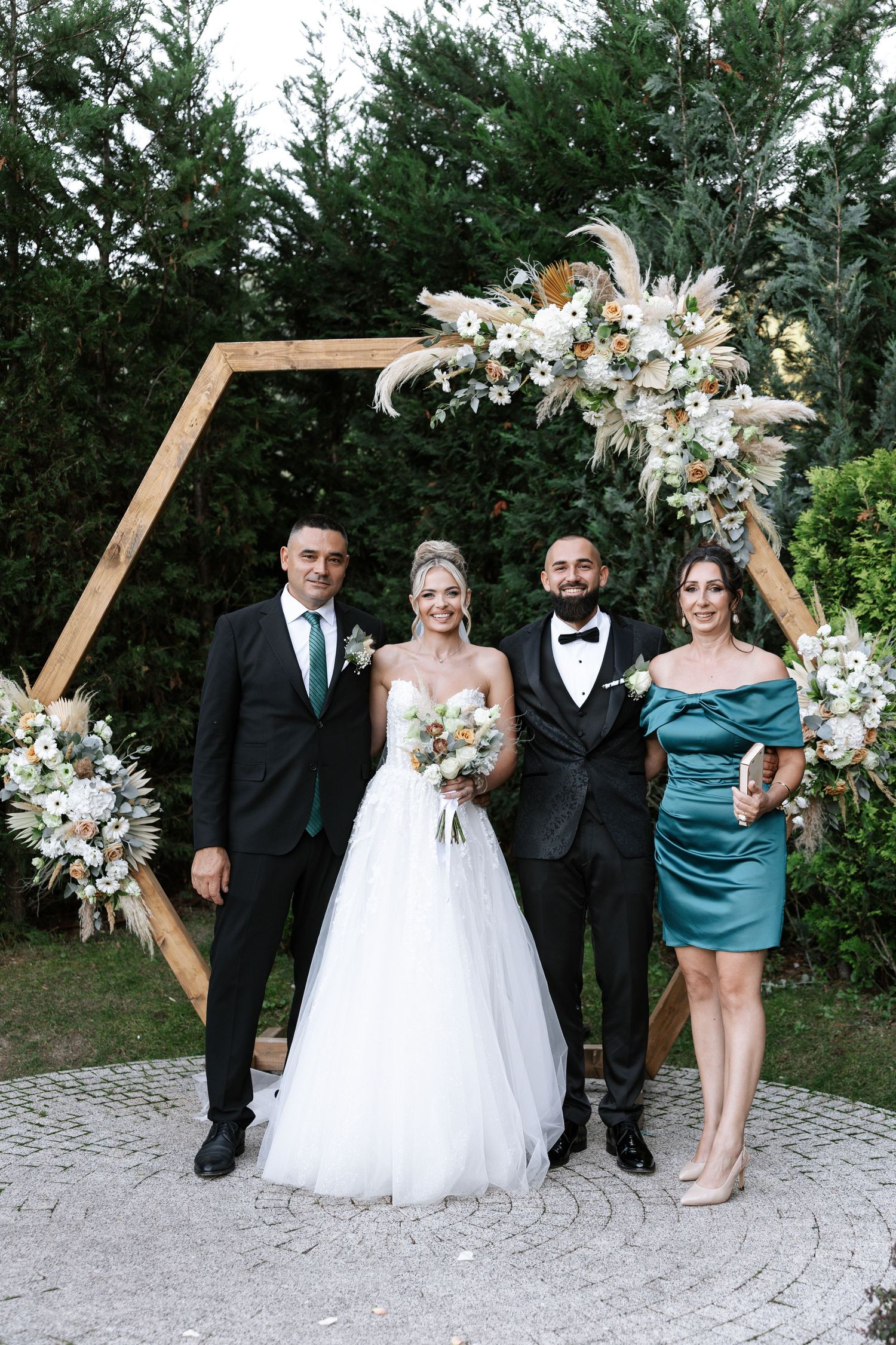 Wedding party poses under hexagonal wooden arch decorated with white floral arrangements and greenery.