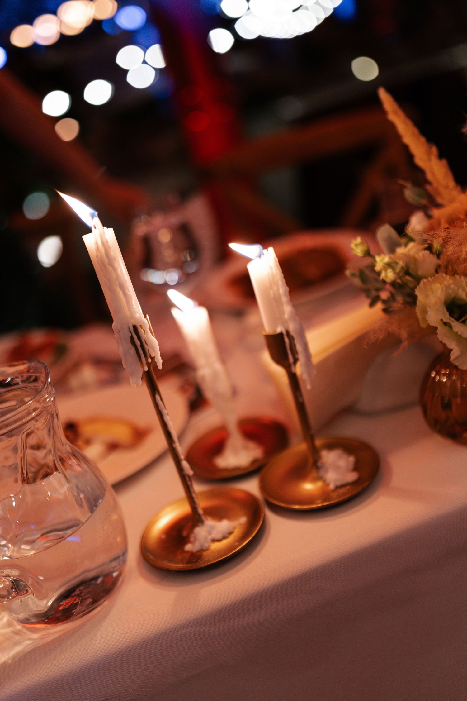 White candles in copper holders illuminate an elegant dinner table setting with glassware and blurred bokeh lights in background.