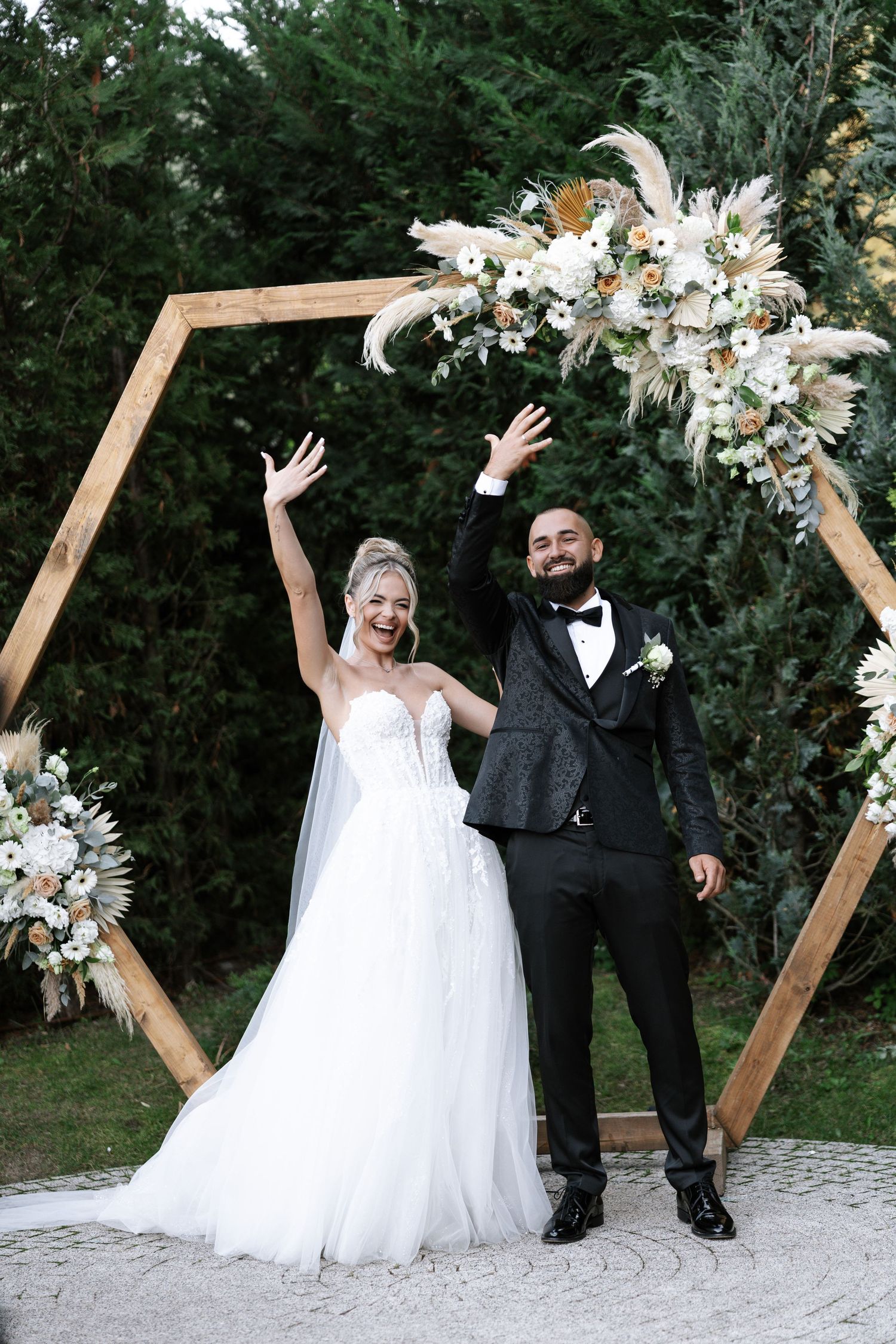 Wedding couple poses playfully in sequence with wooden geometric arch and white floral decorations in outdoor garden setting.