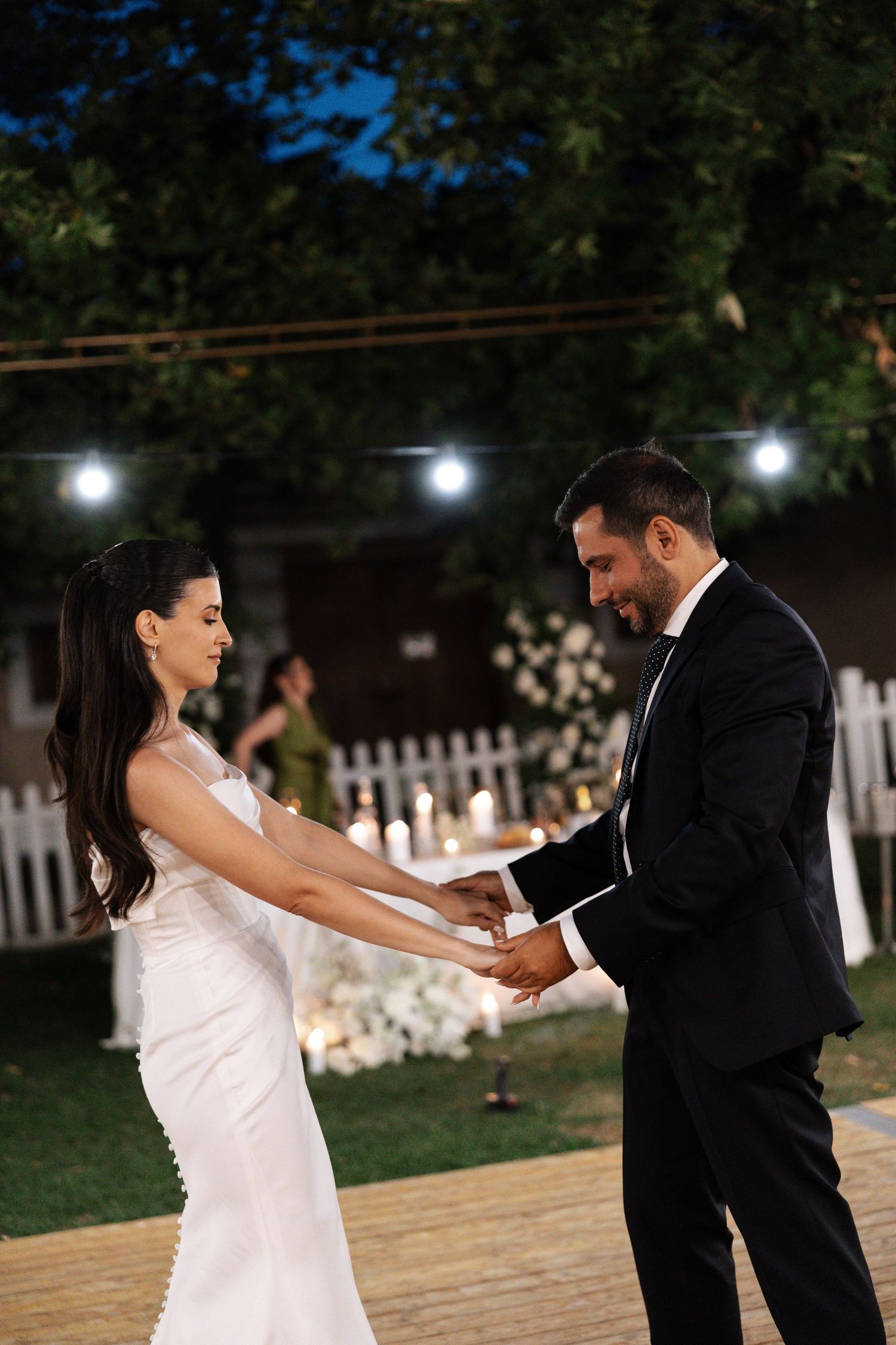 A couple in formal wedding attire dances together on an outdoor wooden dance floor at night.