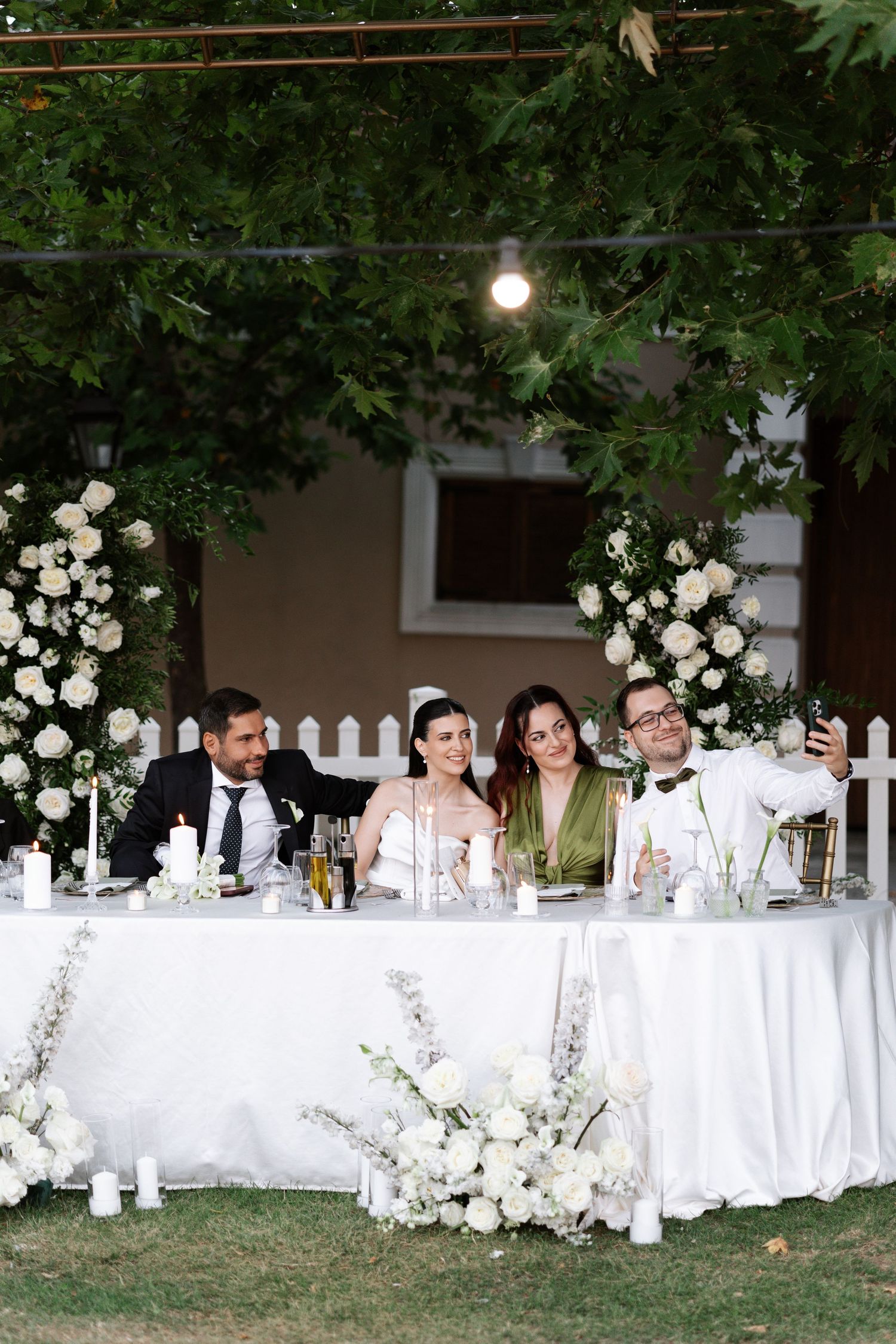 Elegant outdoor wedding reception setup with white tables, floral arrangements and guests seated under trees at dusk.