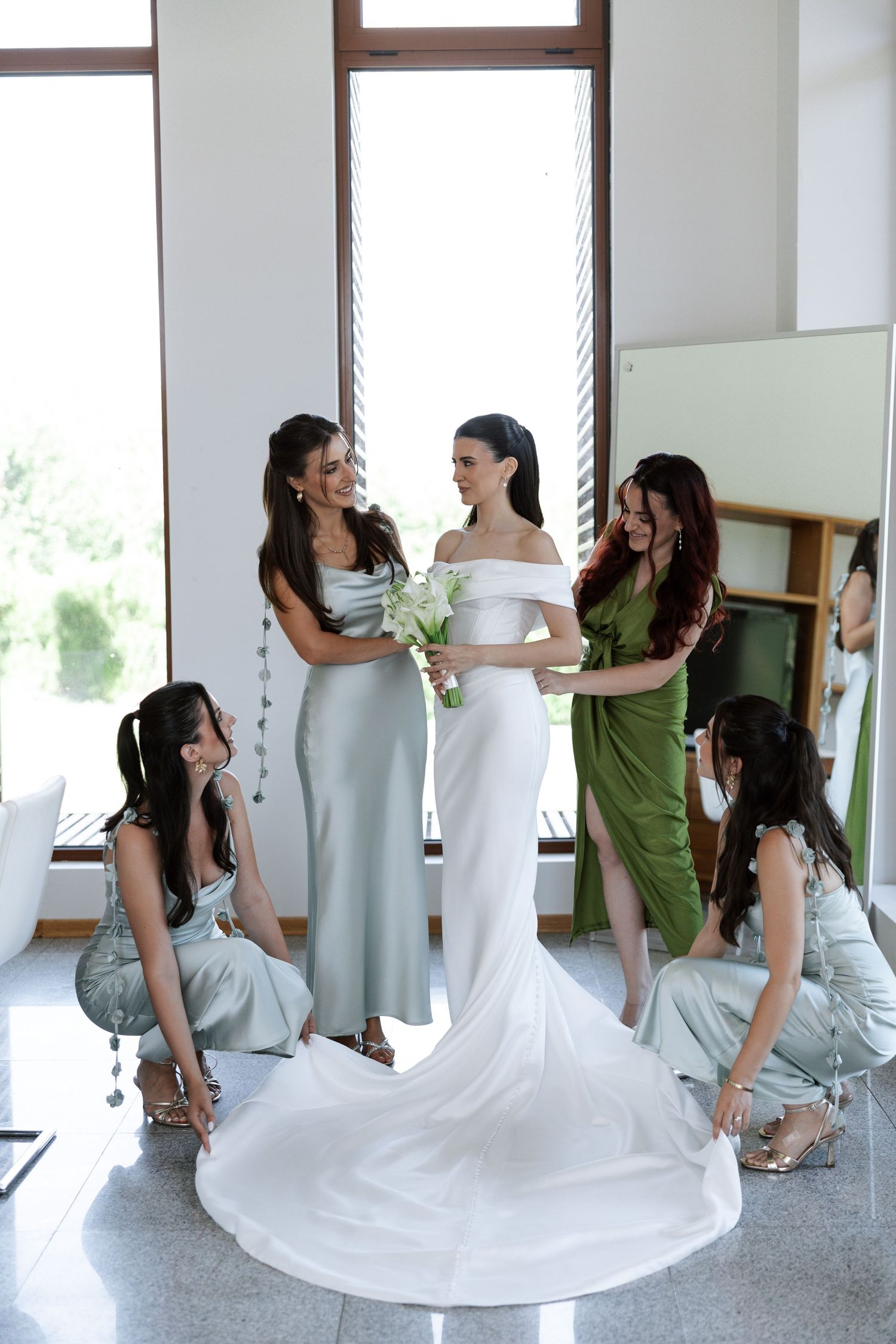 Bridesmaids in light blue dresses gather around bride in elegant white gown with flowing train in bright indoor setting.