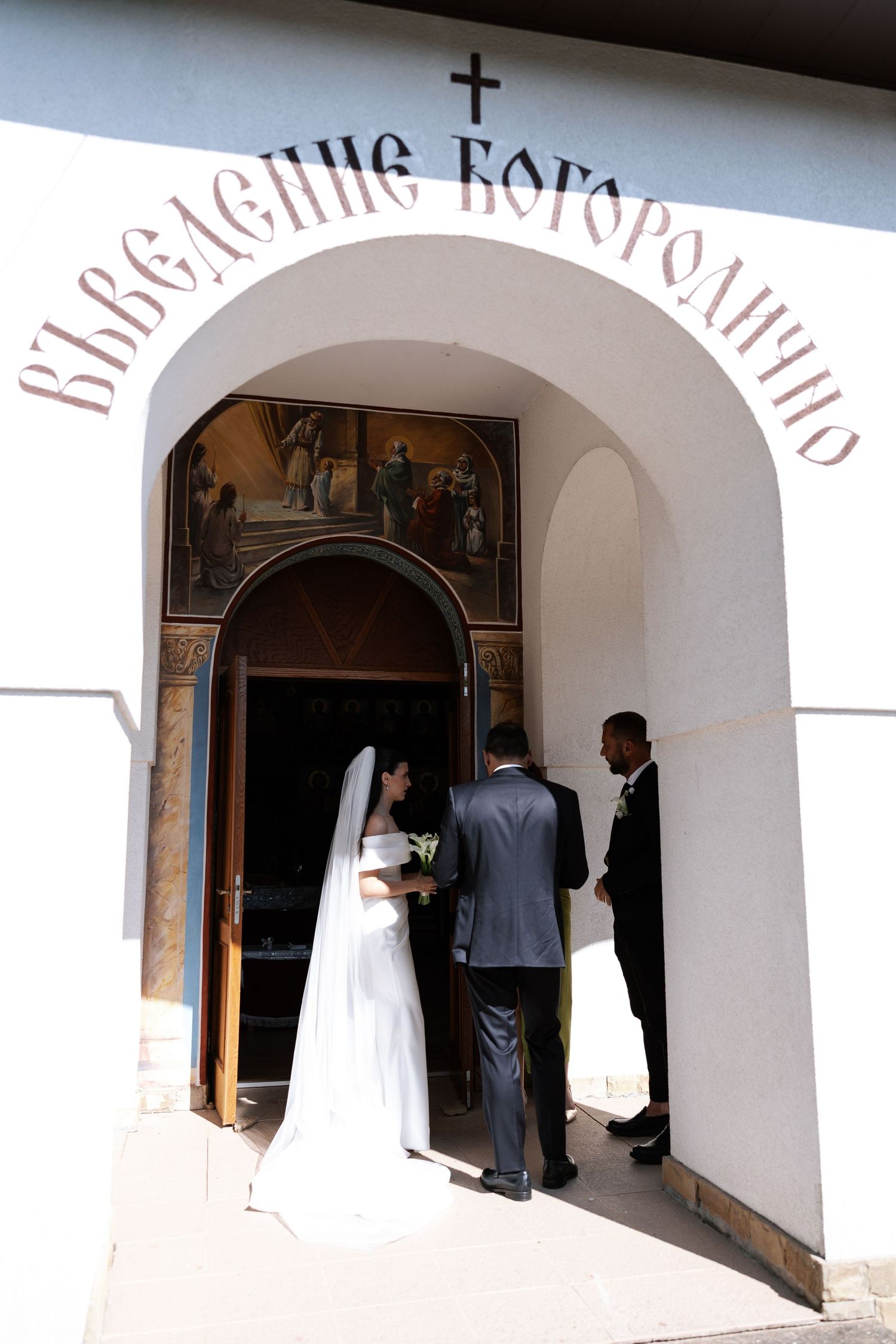 A wedding party enters through an arched church doorway with Cyrillic text above the entrance.