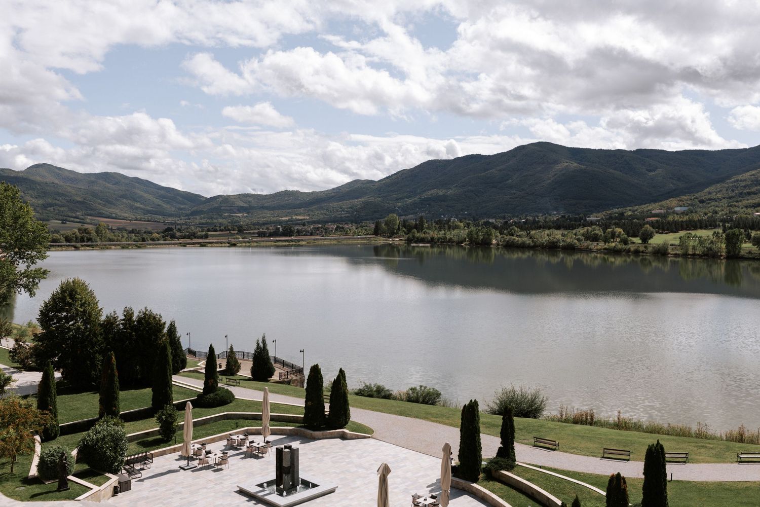Scenic lakeside terrace with manicured cypress trees and mountain views under cloudy skies.