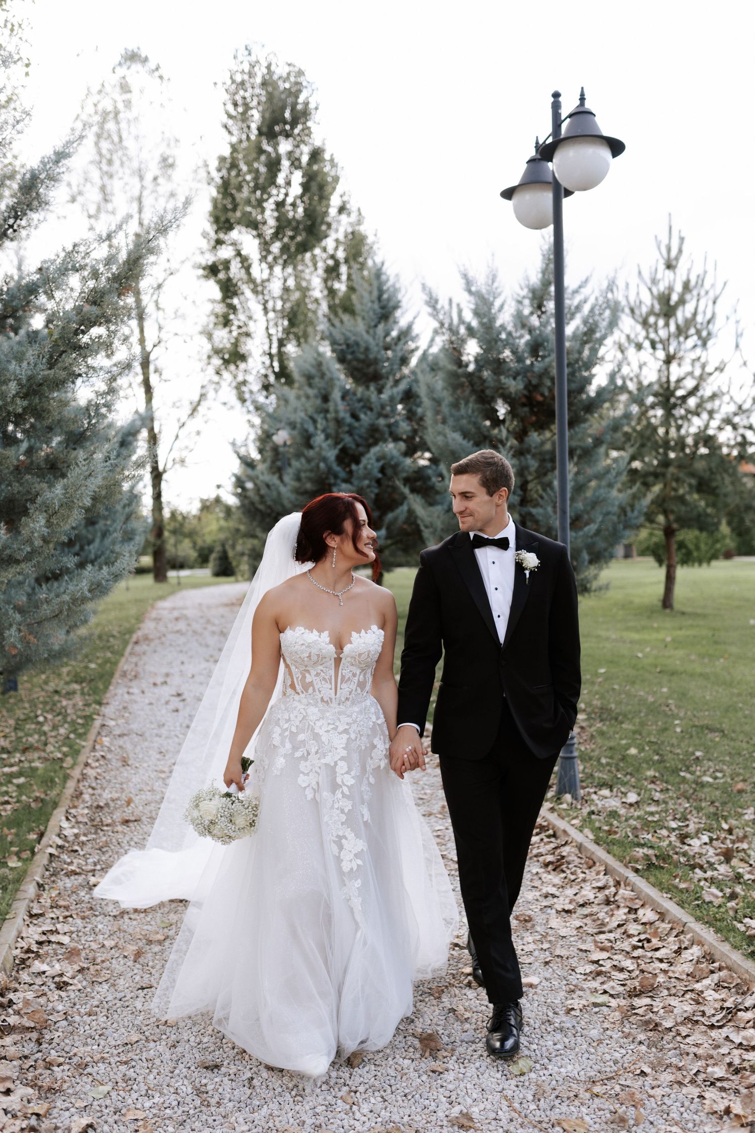 A wedding couple walks hand in hand down a gravel path lined with evergreen trees and a vintage lamppost.