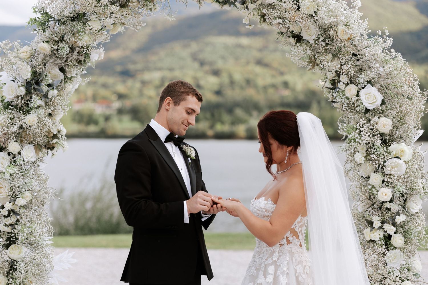 Newlyweds exchange rings beneath ornate white floral arch with mountain landscape.