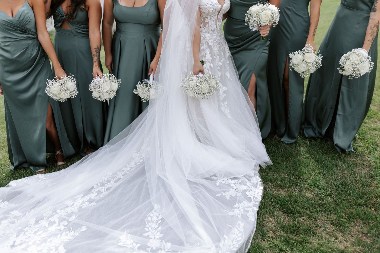 A wedding party with bridesmaids in sage green dresses holding white bouquets next to a bride's flowing lace train.