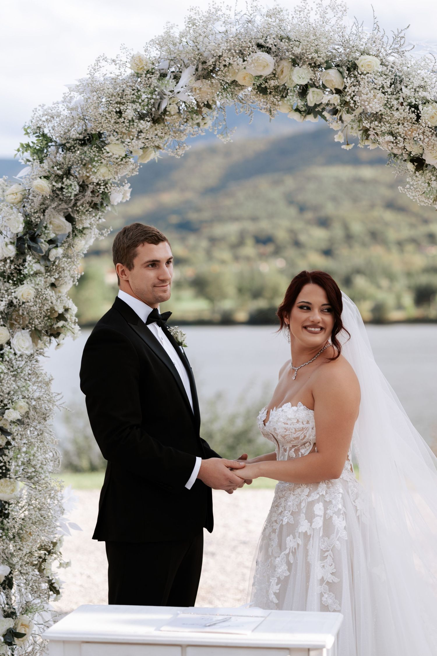 A couple holds hands during their wedding ceremony under a white floral arch.