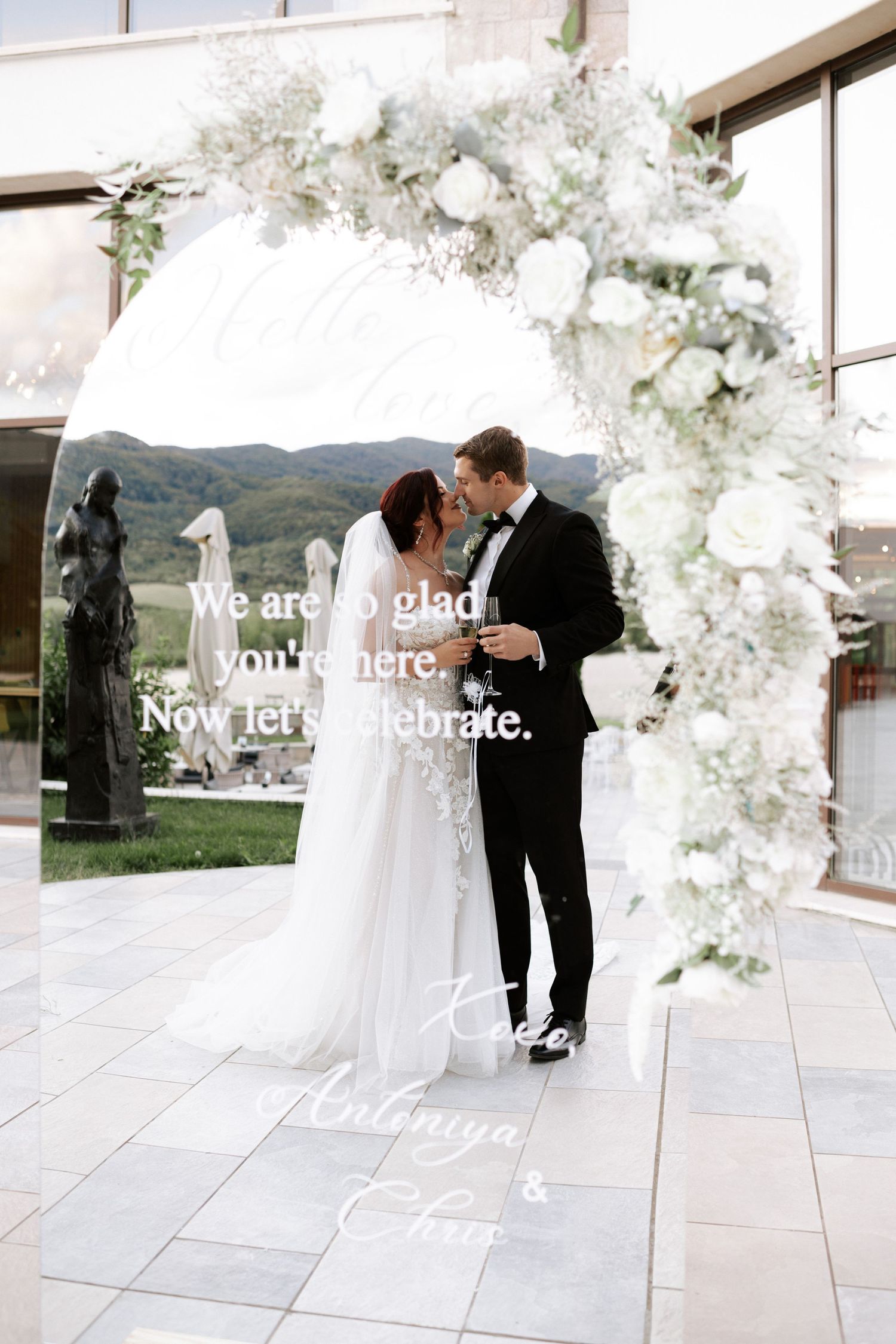 Elegantly decorated white floral arch frames a romantic wedding moment with mountain scenery in the background.