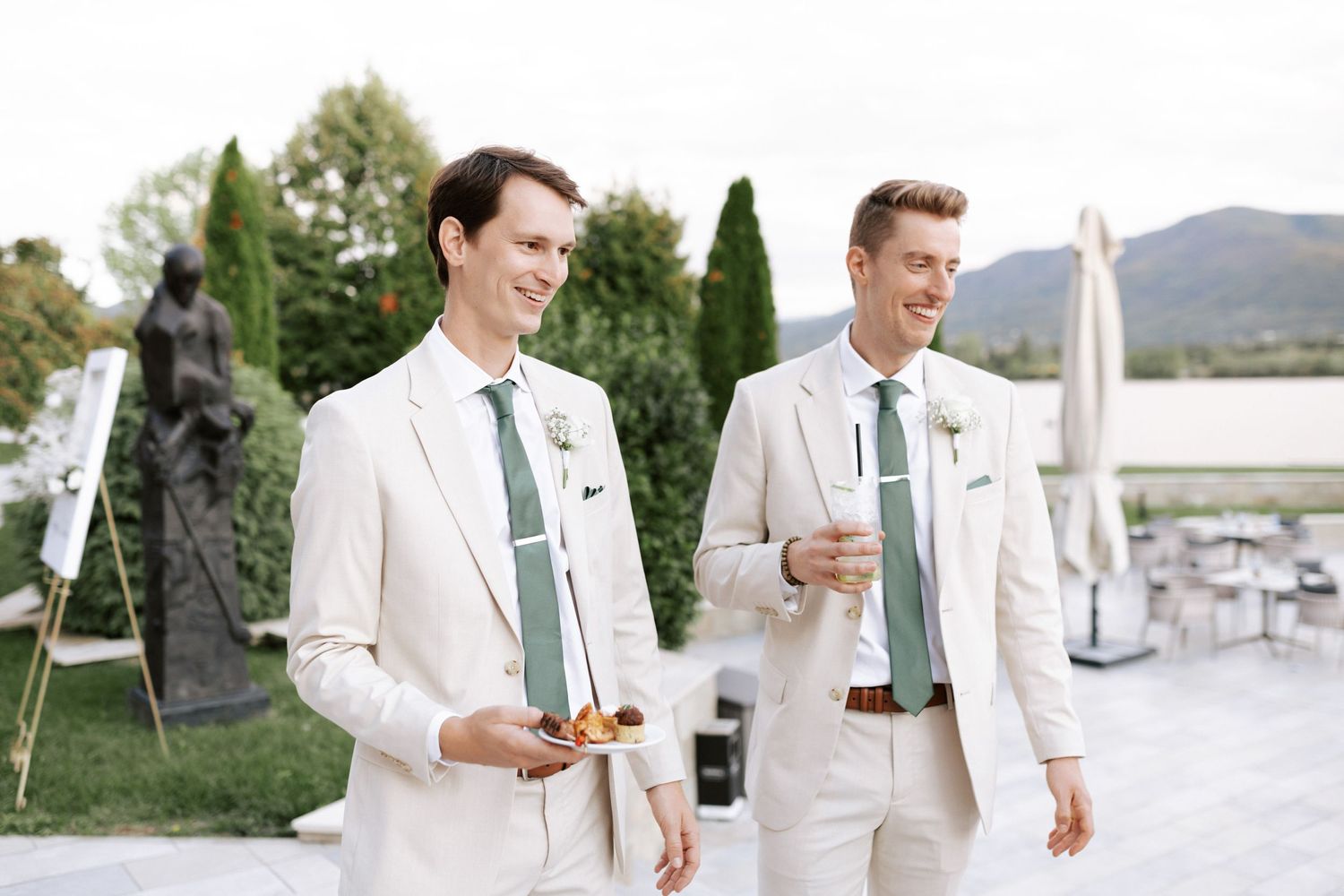 Two groomsmen in cream suits with mint green ties share a laugh outdoors.