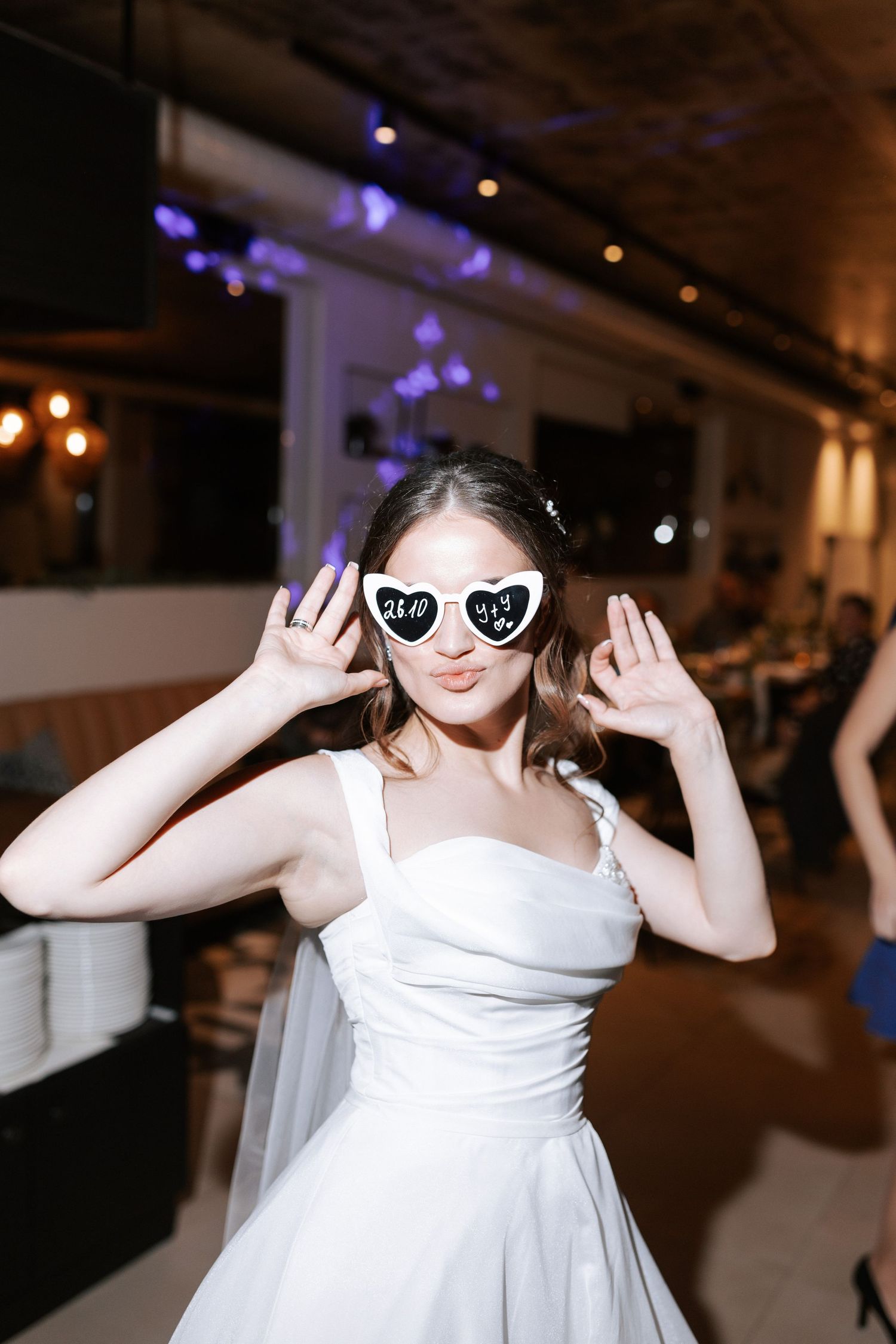 A bride in a white dress poses playfully wearing heart-shaped sunglasses at her wedding reception.