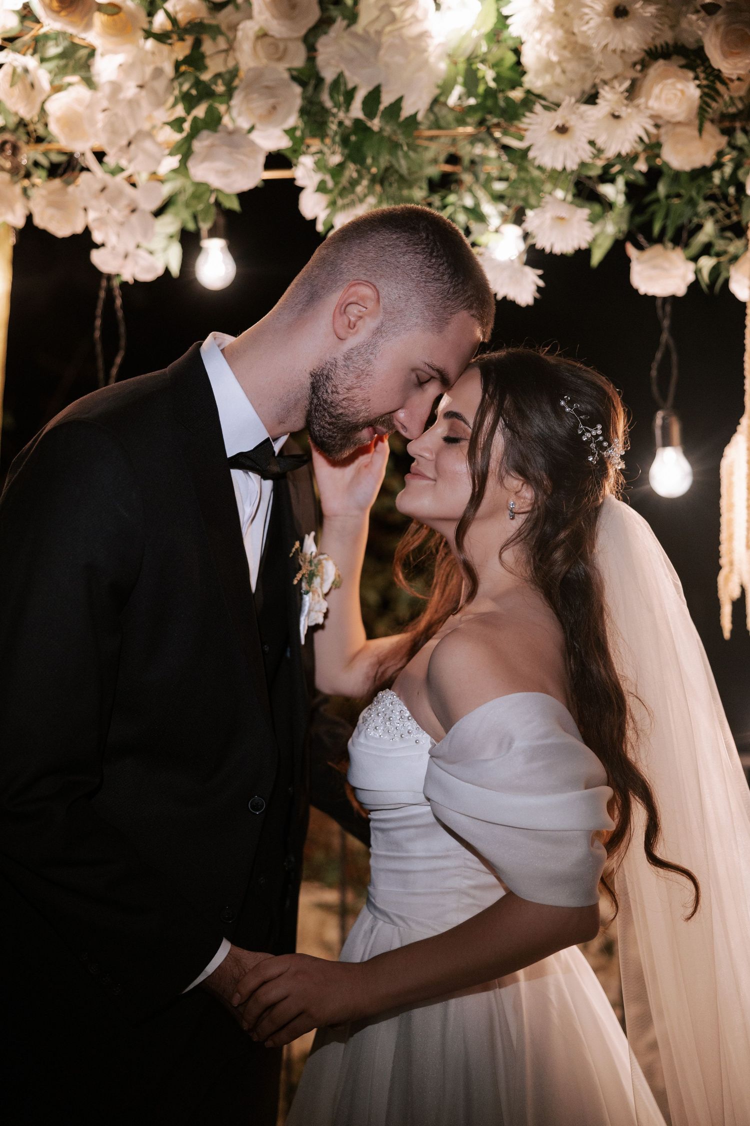 A romantic embrace under hanging white flowers and Edison bulbs at an evening wedding reception.