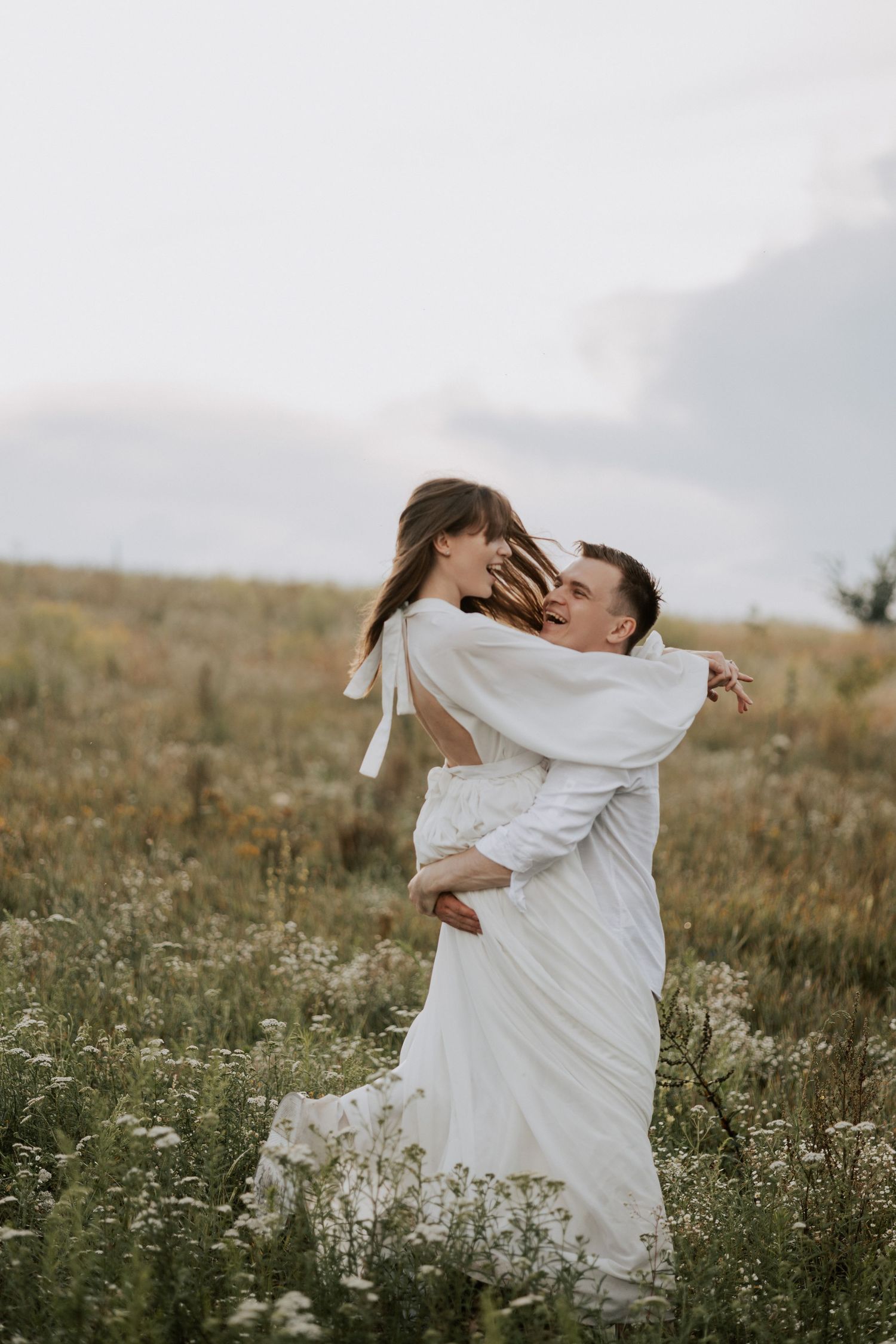 Joyful couple in white embracing in wildflower field during countryside photoshoot.